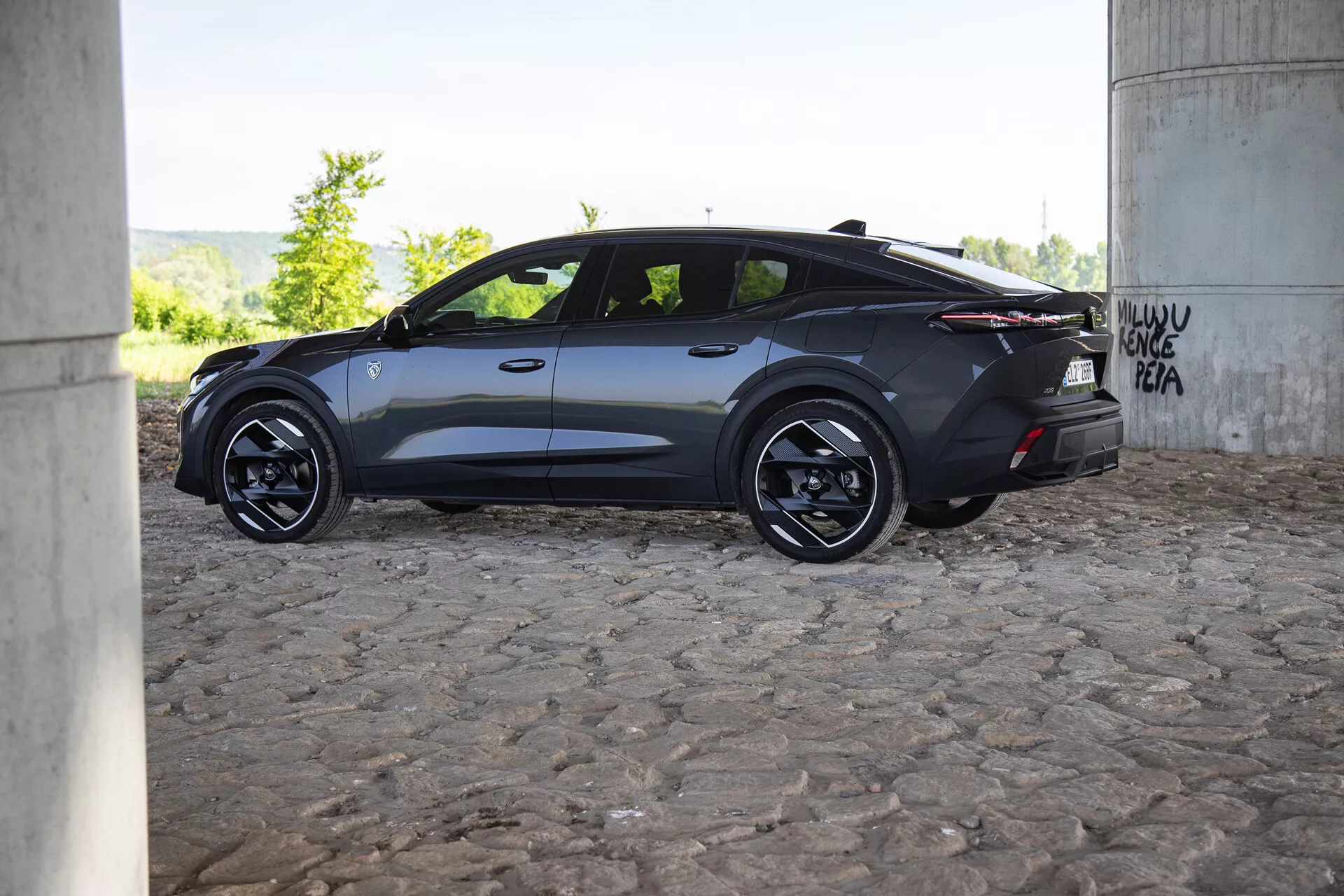 Profilový pohled na moderní tmavě zbarvený vůz Peugeot 408 2023 pod mostem s graffiti, který vyniká aerodynamickým designem, slitinovými koly a tónovanými okny