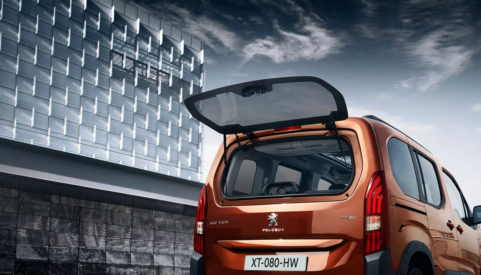 Zadní úhlový pohled na Peugeot Rifter 2018 ukazující jeho hatchbackový design, metalický finiš, otevřené zavazadlové prostor s viditelným nákladovým prostorem, LED zadní světla a městské pozadí.