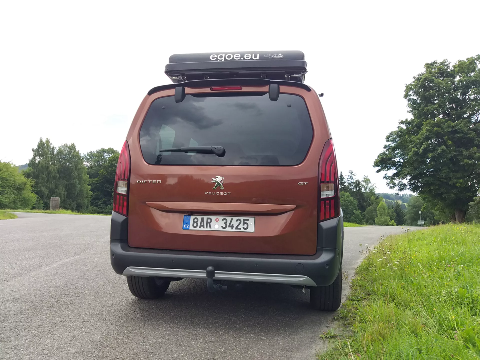 Zadní pohled na Peugeot Rifter 2018, zobrazující zadní okno, koncová světla, nárazník a registrační značku, s viditelným logem Peugeotu a názvem modelu Partner. Auto, parkované proti přírodní kulise, je vybaveno nákladním boxem na střeše a doplněné o příslušenství značky 'egoe.eu', v červené / vínové barvě.