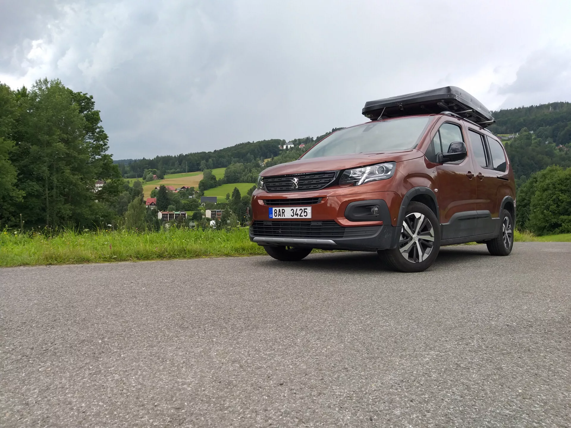 SUV Peugeot Rifter 2018 s kovovým lakem a střešním boxem zachyceno z nízkého úhlu zdůrazňující masku, nárazník a slitinová kola, předvádějící schopnosti pro jízdu v terénu a moderní design včetně LED světlometů a pokročilých systémů asistence řidiče, zaparkované na zpevněné ploše proti venkovské krajině