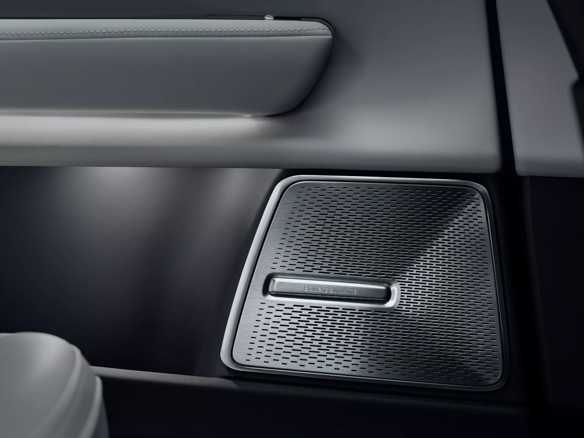Detail interiéru Polestar 3 2023 ukazující vysoce kvalitní mřížku reproduktoru na dveřním panelu auta, s hustým síťováním pro vysokou věrnost zvuku, dvoubarevný design s koženkou se švy a jemně osvětlený kovový povrch odrážející kvalitu audio systému vozidla.