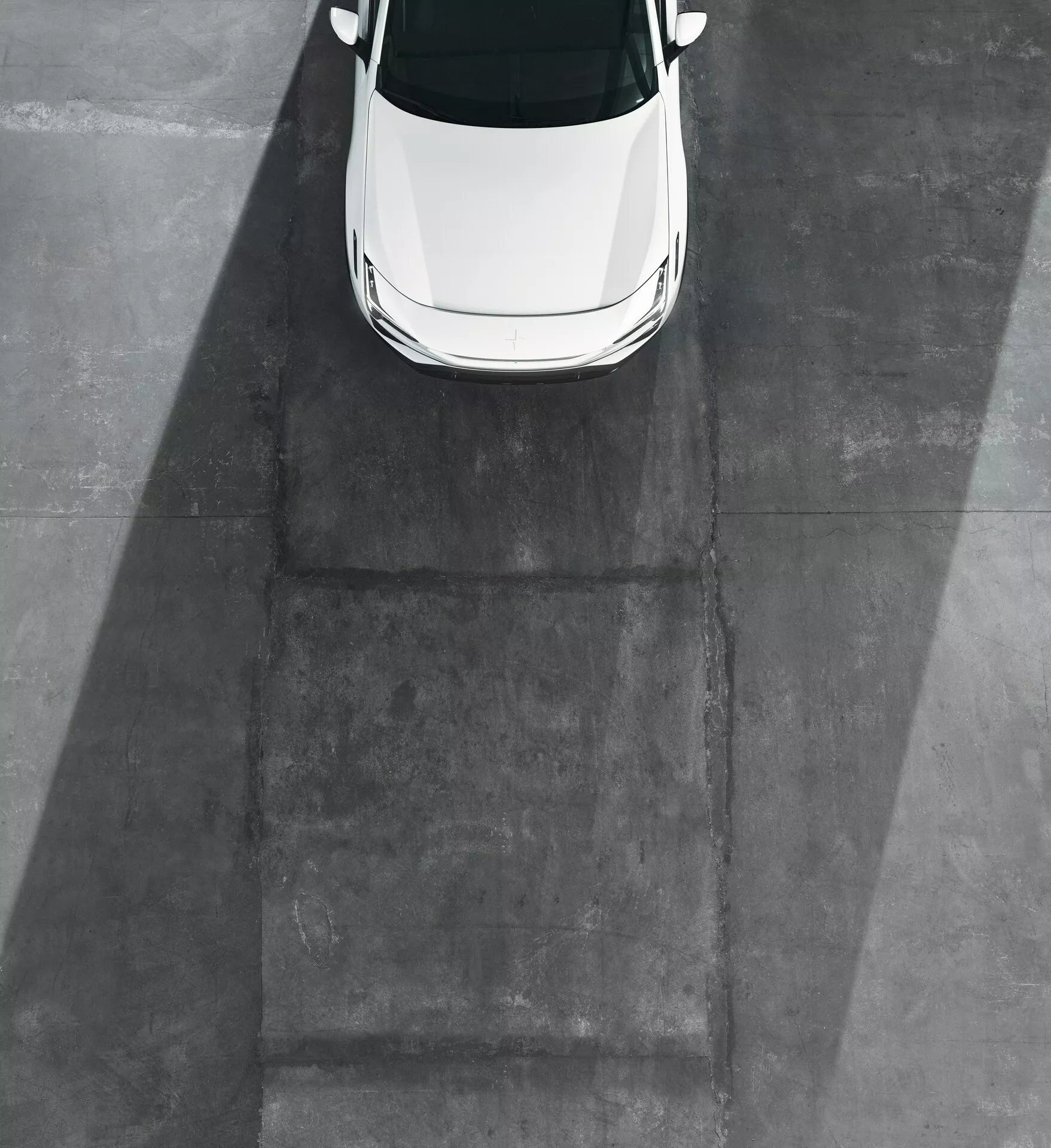 Bílý model Polestar 3 2023 zachycený z ptačí perspektivy s důrazem na střechu, kapotu a část předního čelního skla na šedém betonovém povrchu bez viditelných kol nebo boků, což naznačuje osvětlení brzy ráno nebo pozdě odpoledne