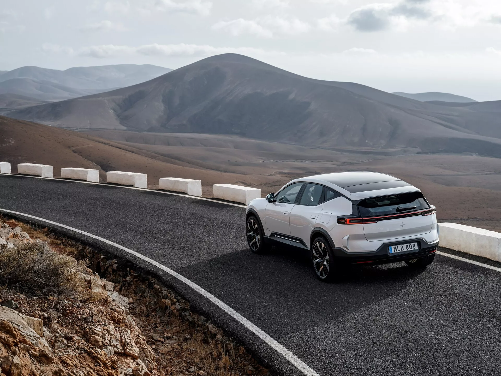 Model Polestar 3 první generace roku 2023 prezentován v dynamickém pohledu ze zadního tříčtvrtečního úhlu na klikaté horské silnici, což zdůrazňuje jeho aerodynamický exteriér, moderní LED světelný pruh a sportovně kupéový design na pozadí malebné horské krajiny, což naznačuje ekologickou jízdu bez viditelné výfukové trubky, což napovídá technologii elektrického nebo hybridního pohonu.