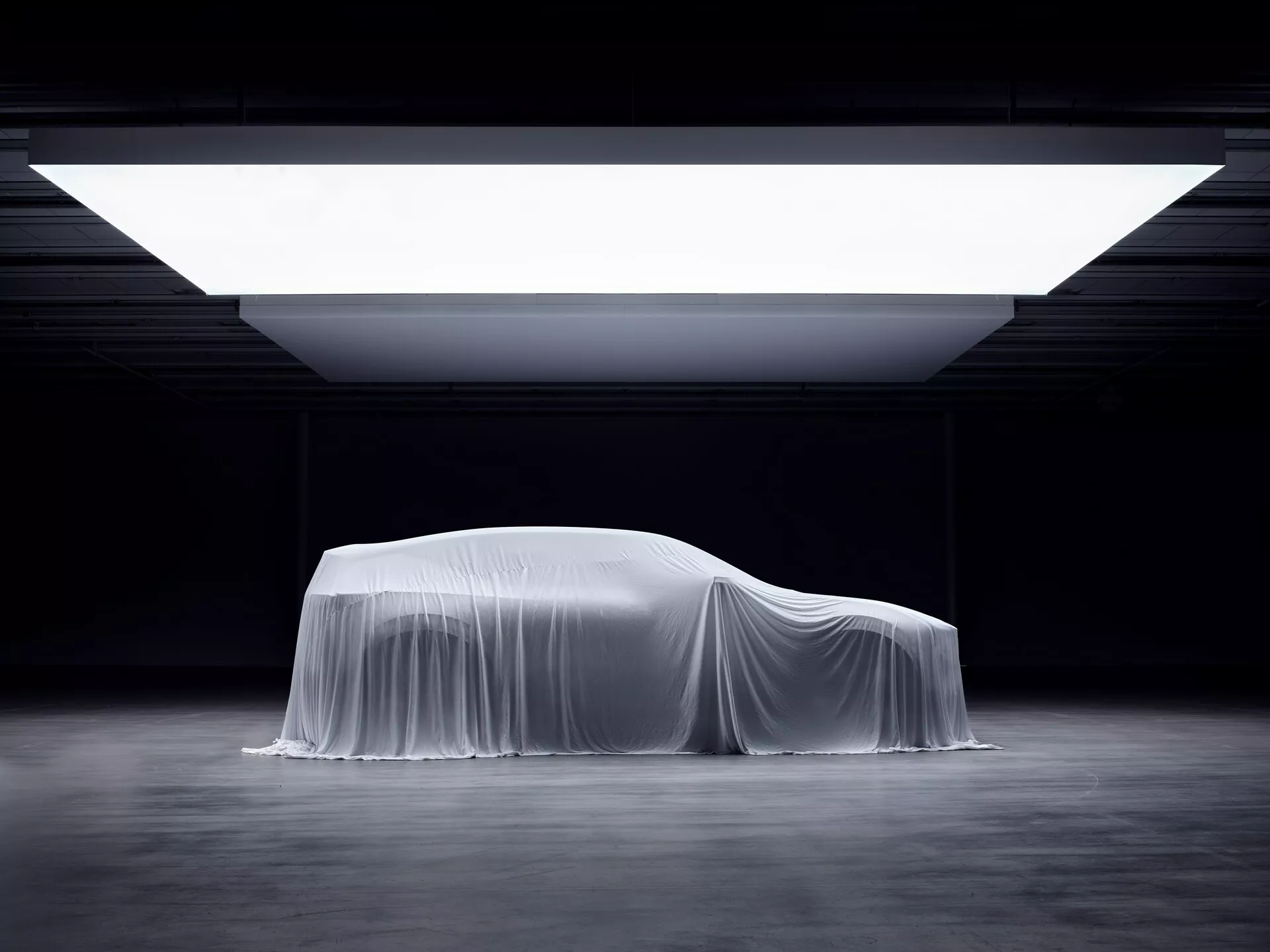 Odhalení první generace Polestar 3 pod plachtou se z profilu zdůrazňující aerodynamický design a kontury kupé pod dramatickým osvětlením showroomu v roce 2023
