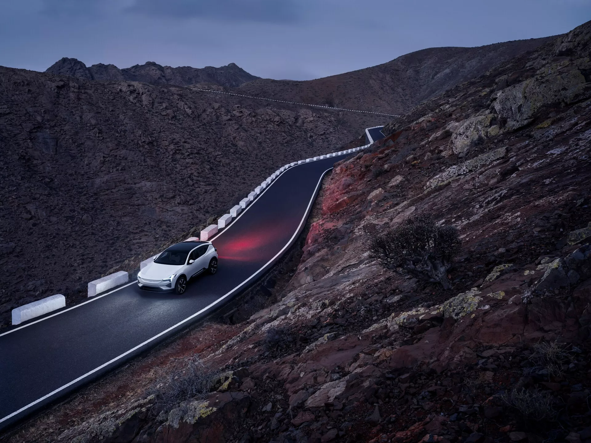 Bílý SUV Polestar 3 2023 jedoucí po klikaté horské silnici za soumraku, předvádějící elegantní design a pokročilé osvětlení