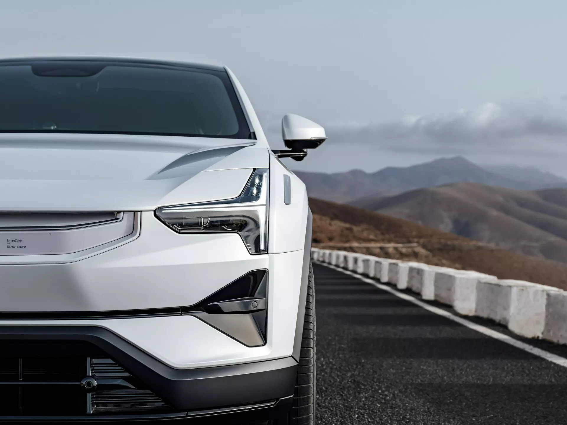 Pohled zepředu zleva na Polestar 3 z roku 2023 prezentující aerodynamickou kapotu, uzavřenou mřížku technologie elektrického vozidla, LED světlomety a elegantní boční zpětné zrcátko na cestě s horským pozadím
