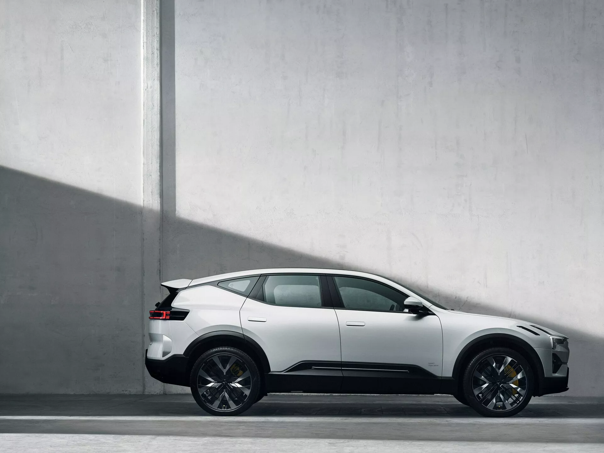 Profilový pohled na SUV Polestar 3 z roku 2023 proti světle šedému pozadí zdůrazňující jeho sportovně kupéovitou siluetu, pětidveřový design, složité vícepaprskové kola a charakteristické linie naznačující pohyb, v přírodních světelných podmínkách naznačující matný lak a moderní, luxusní pocit.