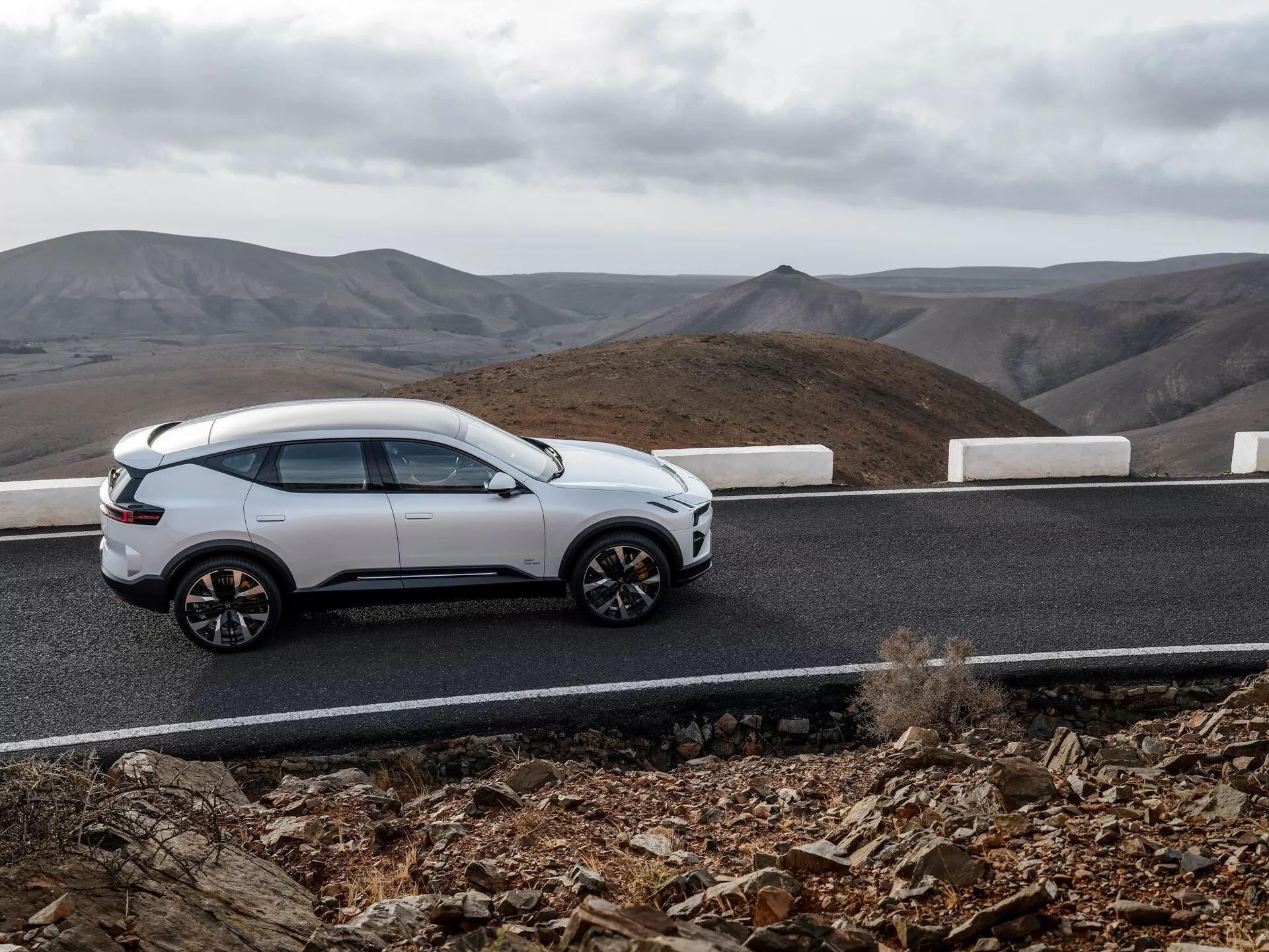 Bílé SUV Polestar 3 ročník 2023 na zakřivené asfaltové cestě s bílým lemem proti pozadí kopcovité krajiny s oblačným nebem, zobrazující boční profil, elegantní design, kola s více paprsky a moderní prvky jako jsou zapuštěné kličky a panoramatické střešní okno, naznačující luxus a všestranné terénní schopnosti