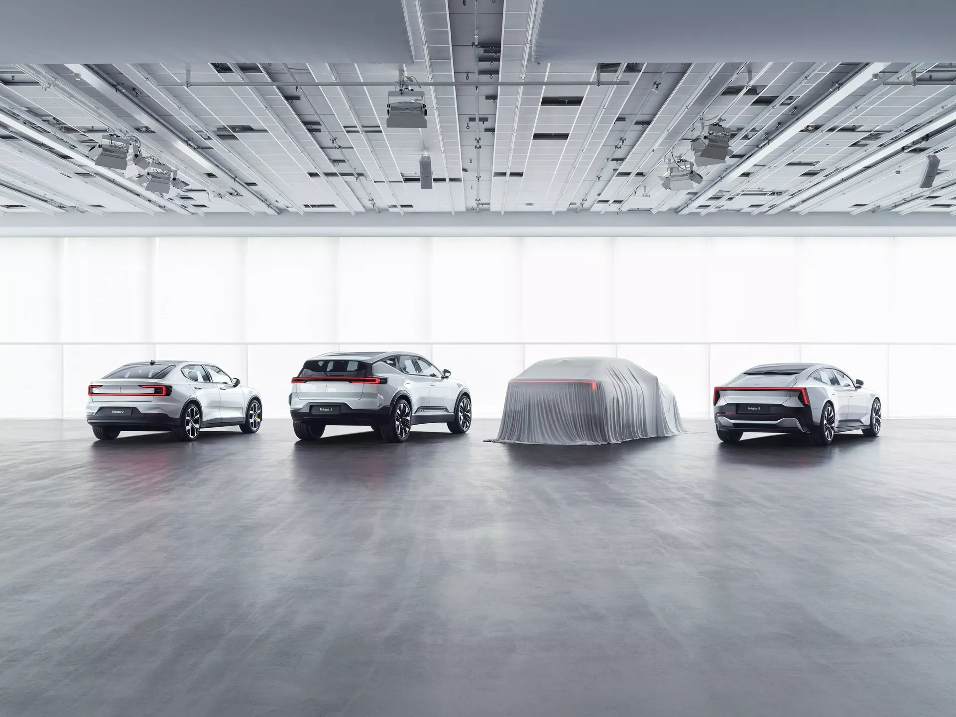 Výstavní sestava Polestar 3 2023 ve showroomu s rozptýleným osvětlením, zobrazující profilové a čtvrtinové pohledy na tři viditelná auta a jeden krytý model naznačující odhalení, znázorňující pokročilé technologické funkce