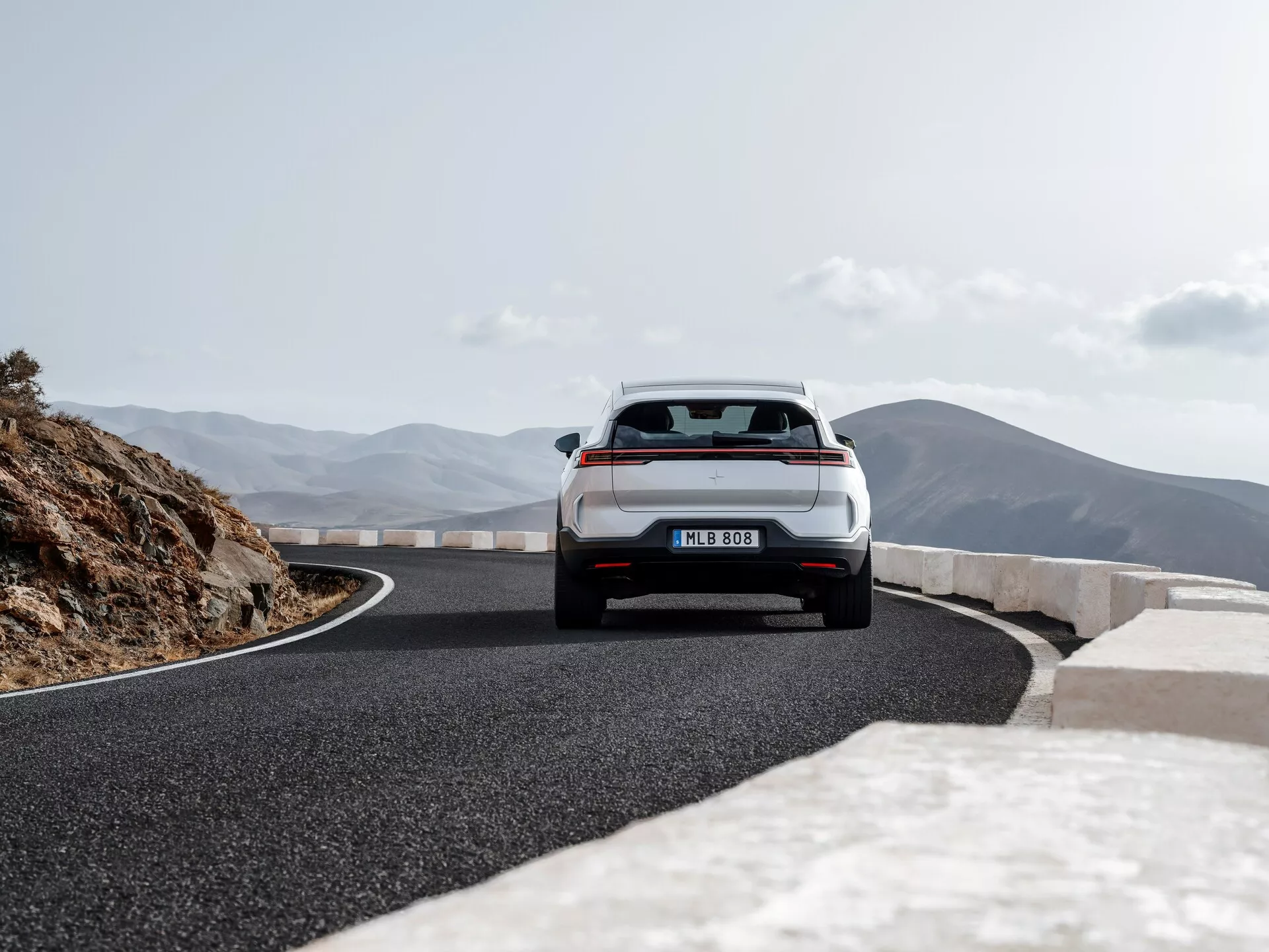 Polestar 3 první generace roku 2023 zachycený z zadní perspektivy ukazuje celou zadní část včetně kufru, zadního nárazníku, zadních světel a části zadní střechy na malebné horské silnici, zdůrazňuje jeho hatchback nebo crossover SUV design s LED zadními světly, dvojitým výfukovým systémem a stěračem zadního okna pro zvýšenou bezpečnost a výkonnostní schopnosti