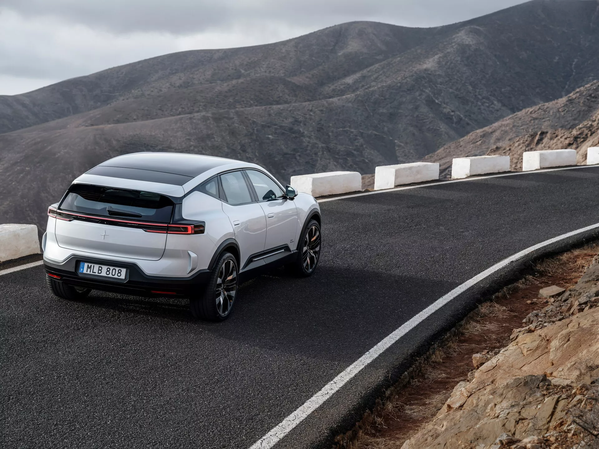 Bílé SUV crossover Polestar 3 první generace 2023 ukazuje elegantní, sportovní design s velkými slitinovými koly a charakteristickými LED zadními světly, zaparkované na klikaté horské cestě, z pohledu ze zadní části a boku zdůrazňující jeho moderní linie a technologické vlastnosti.