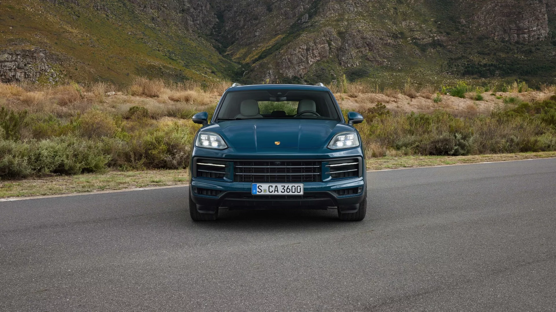 Porsche Cayenne 2023 (9YA Facelift) z pohledu přímo z očí ukazuje LED světlomety, výraznou masku s logem značky, sportovní design kapoty a integrované parkovací senzory na pozadí horské krajiny, což naznačuje luxus a dobrodružství