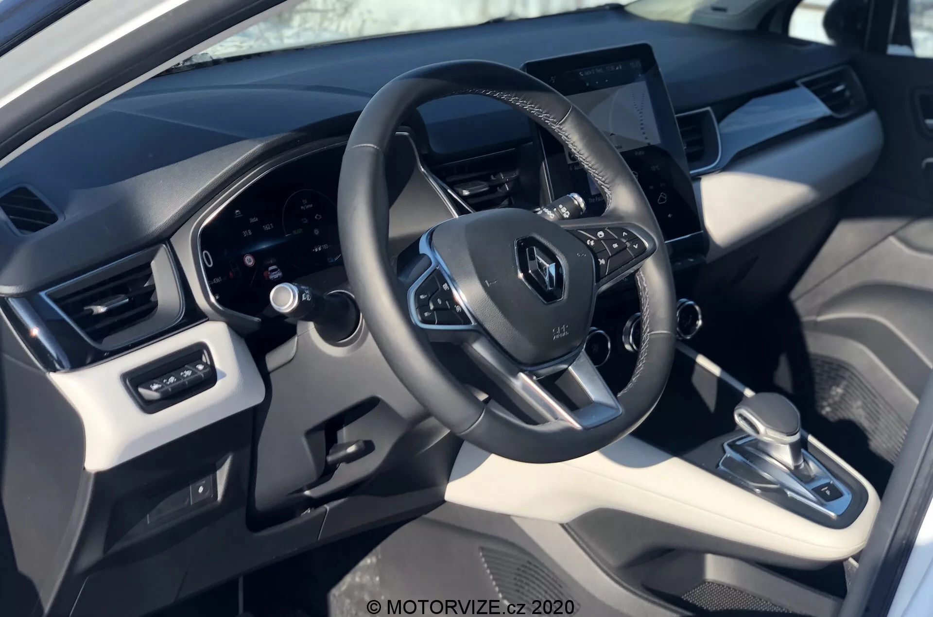 Interiér vozu Renault Captur 2020 z pohledu řidiče, zobrazující volant s logem Renault, ovládací tlačítka, digitální přístrojový panel a centrálně umístěný dotykový infotainment systém. Snímek zdůrazňuje směs syntetické kůže a plastových materiálů s kovovými akcenty, s automatickou převodovkou, systémem klimatizace a ovládáním elektrických oken a zrcátek.