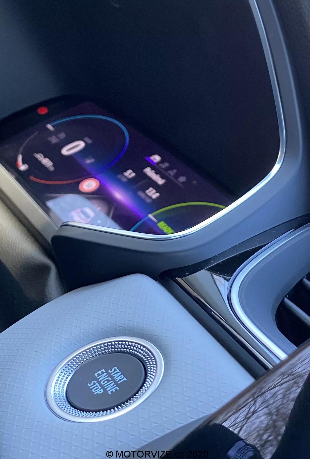 Interiér Renaultu Captur 2020 se zaměřením na středovou konzoli a přístrojovou desku z perspektivy předního sedadla. Živý infotainment displej zobrazuje barevné rozhraní naznačující pokročilé multimediální funkce, včetně navigace, přehrávání médií a připojení. Pod displejem se nachází kruhový ovladač s chromovaným povrchem označený 'Engine Start Stop', který zdůrazňuje bezklíčový startovací systém. Středová konzole je zakončena kvalitním strukturovaným povrchem, který zlepšuje atmosféru interiéru. Vlevo je částečně viditelný držák na nápoje nebo úložný prostor.
