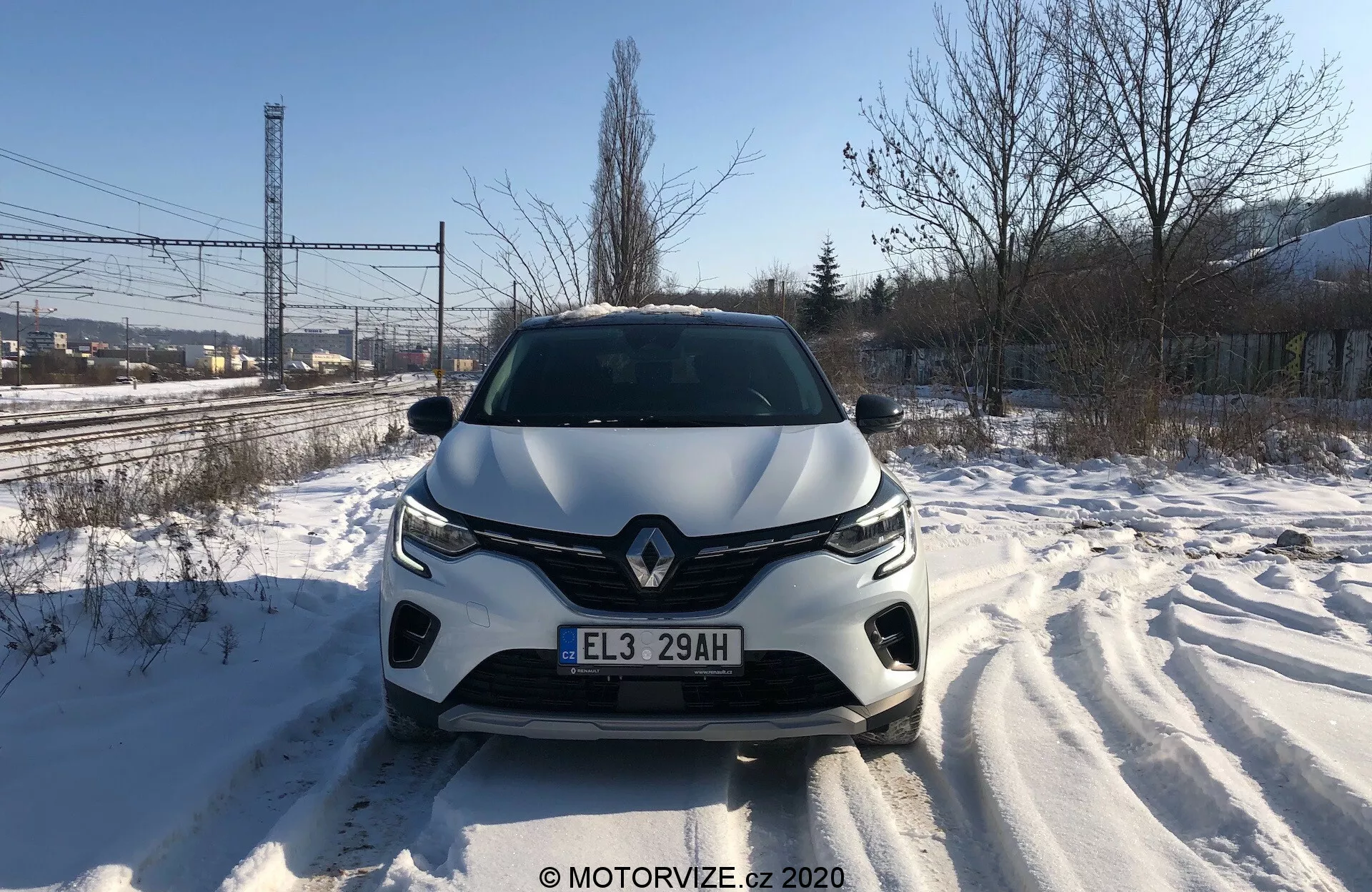 Přímý přední pohled na Renault Captur 2020 na sněhem pokryté silnici, ukazující detailně přední mřížku, světlomety, tvarovanou kapotu a přední nárazník během dne. Viditelné stopy pneumatik svědčí o nedávném pohybu. Design vozu naznačuje aerodynamické tvary, s výrazným logem značky a pokročilým osvětlovacím systémem. V předním nárazníku mohou být integrovány senzory nebo kamery asistence pro řidiče.