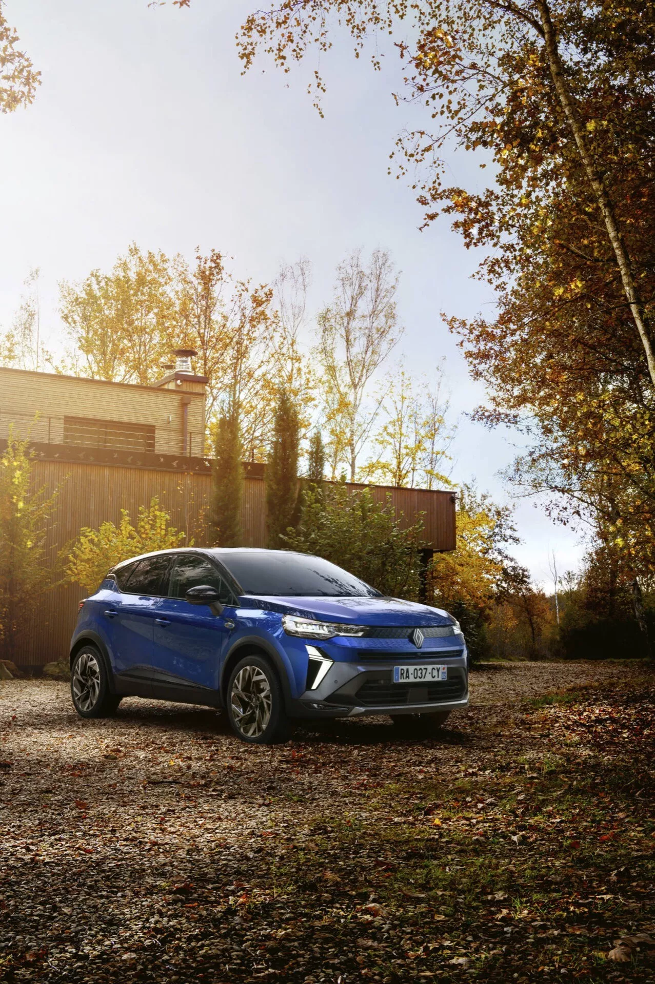 Model Renault Captur II facelift 2024, modré kompaktní SUV zaparkované venku v prostředí podzimního listí, ukazující dominantní a úhlový pohled z přední levé strany s detailními prvky designu, jako jsou ostré linie, výrazná světla a slitinová kola, zdůrazňující potenciální prvky jako panoramatická střecha a moderní technologie v podzimním prostředí