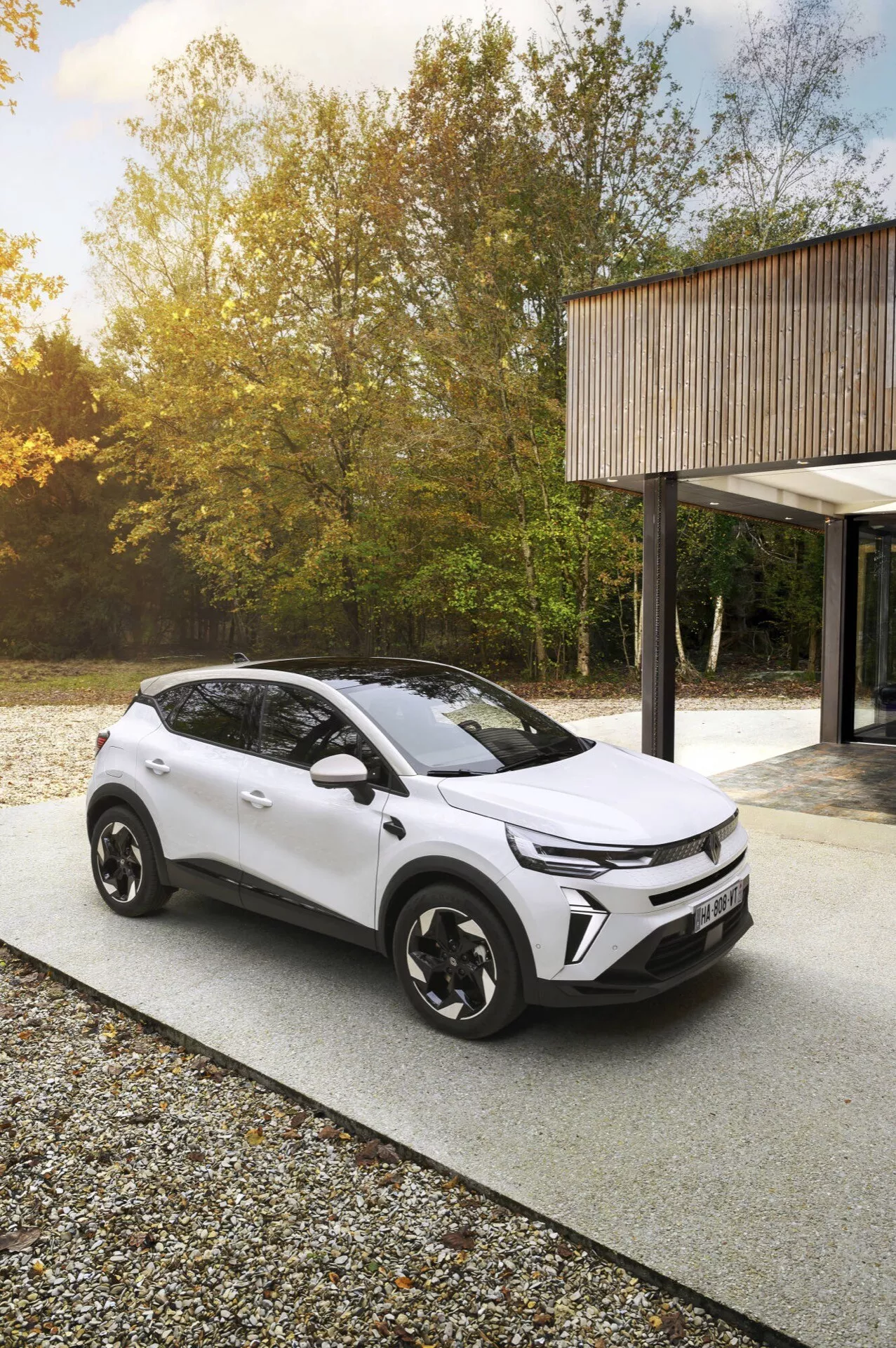 Facelift Renault Captur 2024 zachycen z vysokého úhlu v exteriéru, možná v lesnatém prostředí, zdůrazňuje moderní design vozidla a technologické prvky, jako jsou LED světlomety, dvoubarevné barevné schéma a aerodynamická kola, naznačující ekologicky šetrný elektrický nebo hybridní model.
