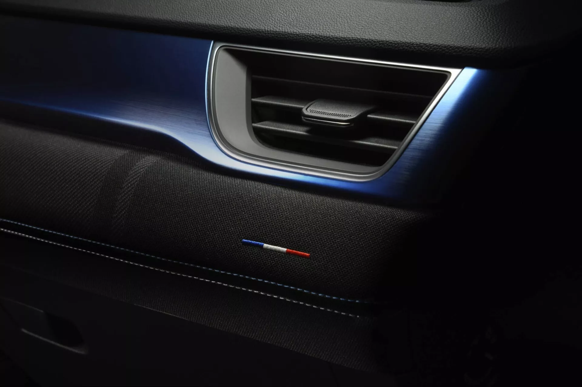 Detail interiéru Renault Captur II facelift 2024 zobrazující dveřní panel s elegantní kovovou klikou, jemné modré ambientní osvětlení, prémiovou vložku dveří se švy a emblémem národní vlajky naznačující speciální výbavu nebo sportovní balíček
