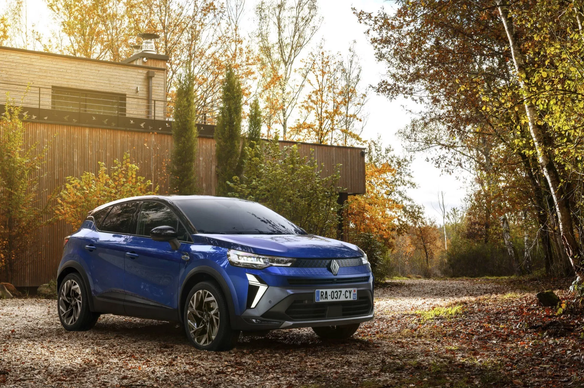 Modrá Renault Captur II facelift 2024, kompaktní SUV zaparkované venku s výhledem na pravou přední část, zaměřené na její moderní designové linie, výraznou přední mřížku, LED světlomety a slitinová kola, na pozadí podzimní krajiny pod přirozeným ranním nebo pozdním odpoledním sluncem, které zdůrazňuje její estetickou přitažlivost a technologický pokrok