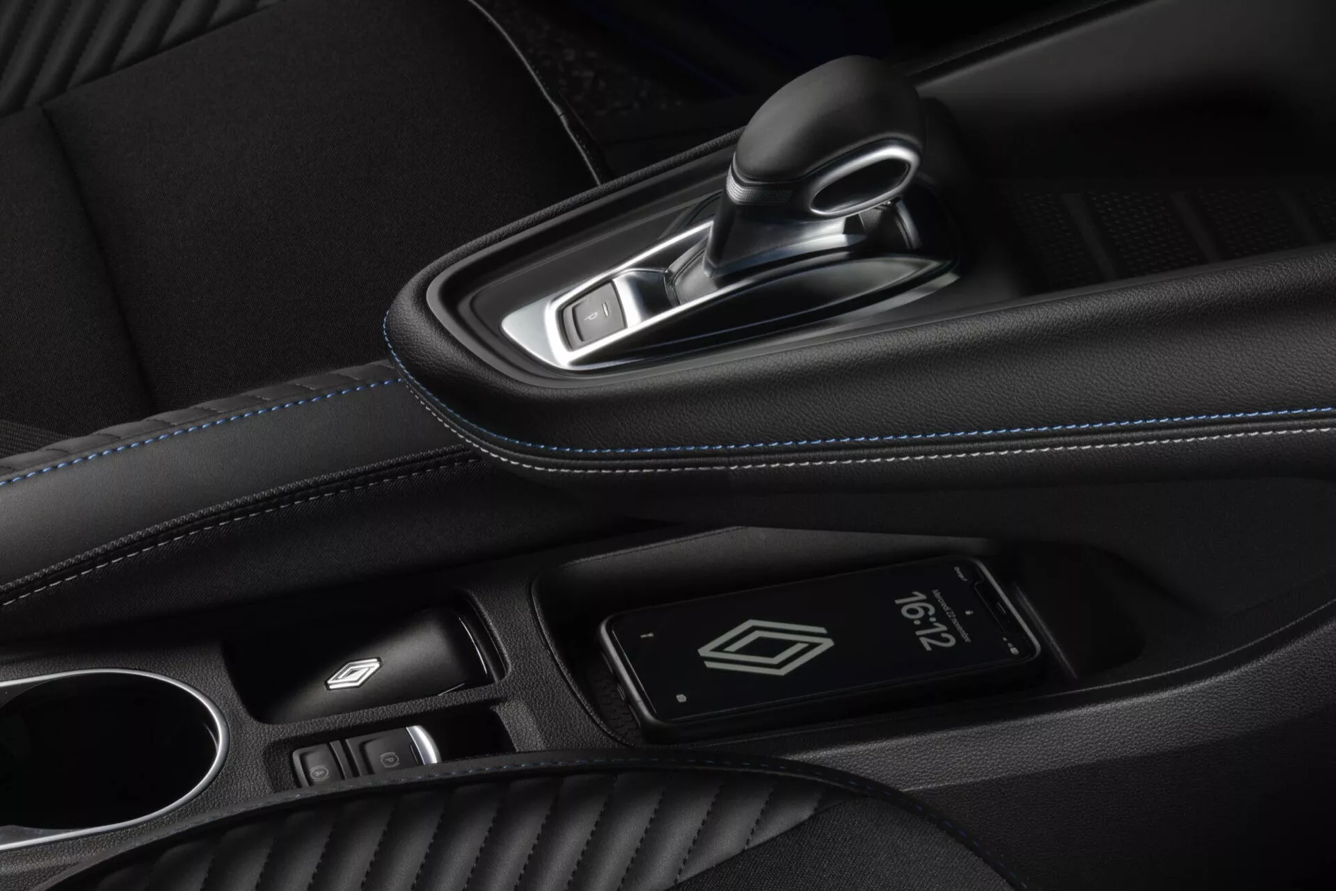 Interiér Renault Captur II facelift 2024 se zaměřením na středovou konzoli a přední kabinu, včetně kožené řadicí páky s chromovanými akcenty, tlačítka pro bezklíčové startování, ovladače dotykového panelu pro informačně-zábavní systém a kožené loketní opěrky s modrým prošíváním z vysokého úhlu směrem k straně spolujezdce