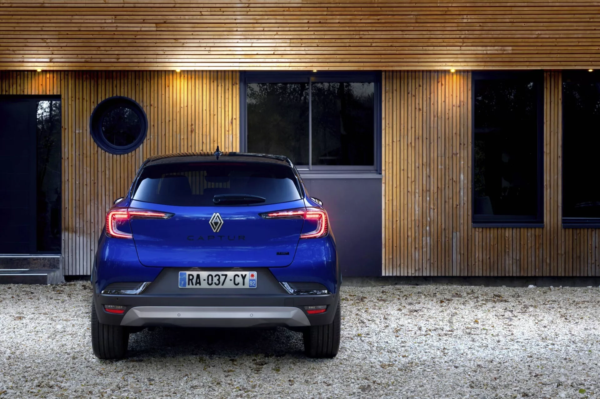 Zadní pohled na modrý Renault Captur (II Facelift) rok 2024 s LED zadními světly, parkovaný před moderním obytným domem, s parkovacími senzory, dvojitým výfukem a anténou ve tvaru žraločí ploutve
