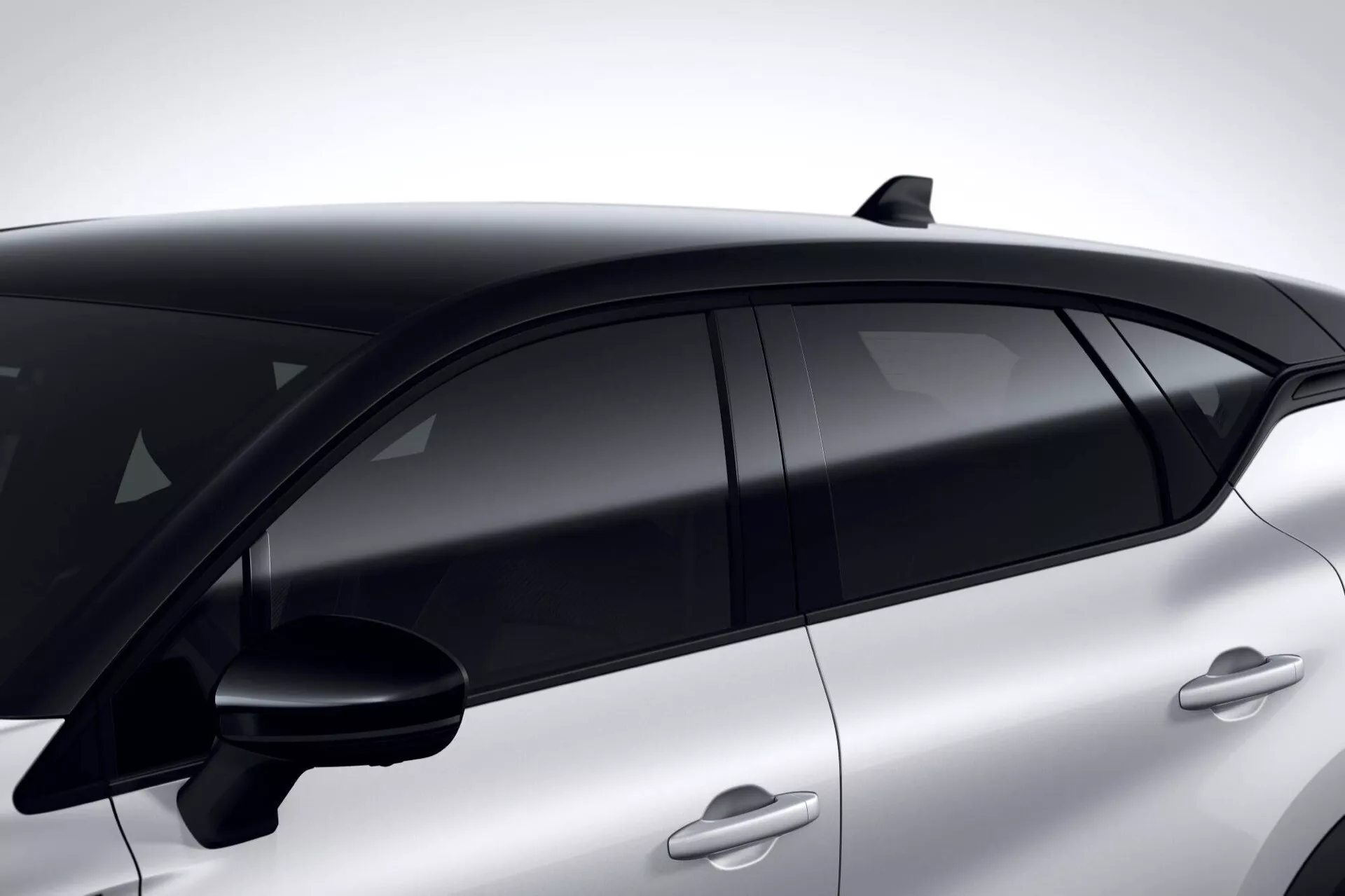 Detailní boční pohled na Renault Captur 2024 (II-facelift) zvýrazňující jeho aerodynamickou střechu, sofistikovanou žraloční ploutev anténu a moderní design oken s černě tónovaným, bezrámovým sklem ohraničeným černými B-sloupky, umístěný proti elegantnímu, sportovnímu profilu s barvou sladěnými, zapuštěnými klikami dveří naznačujícími pokročilou technologii bezklíčového vstupu