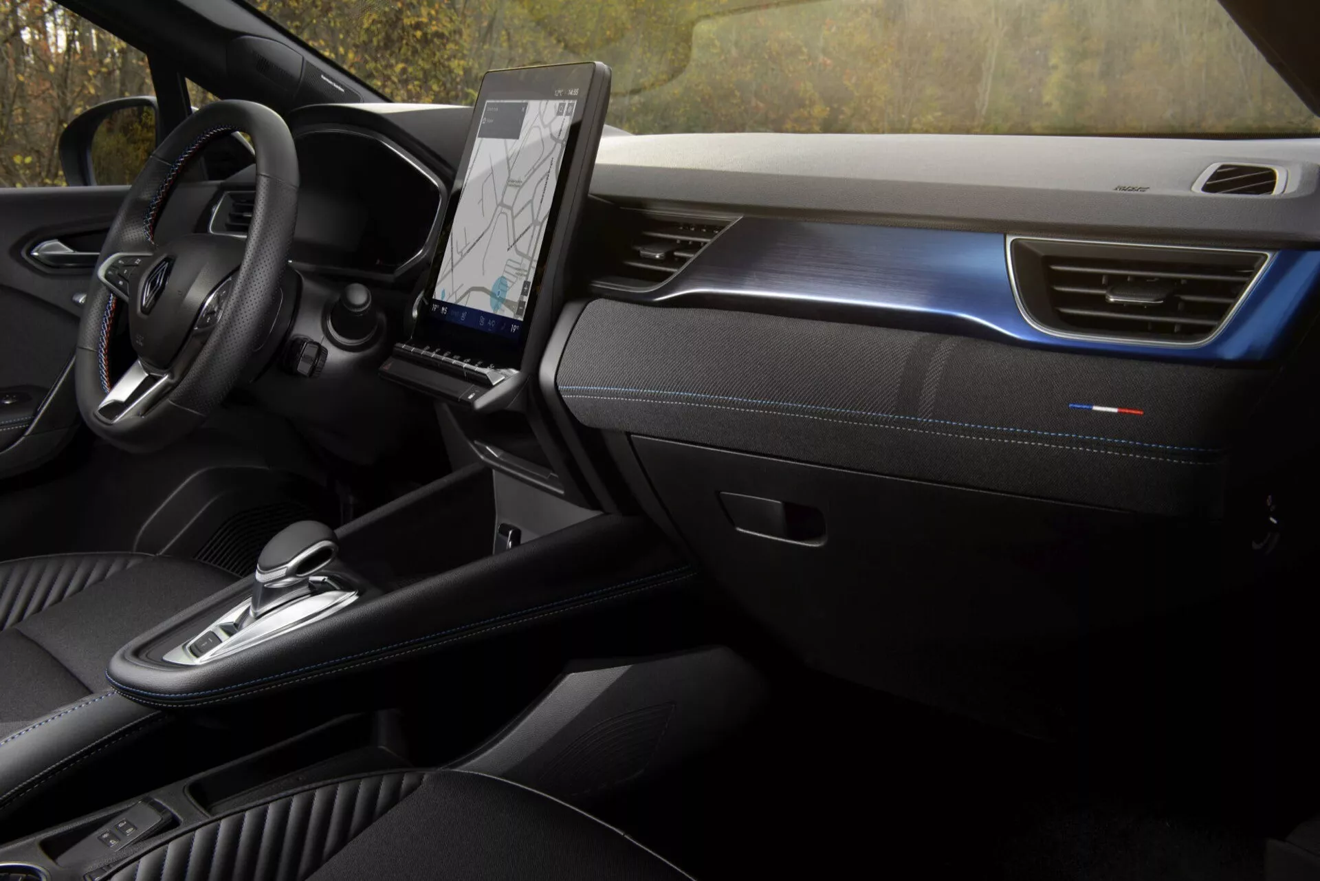 Interiér Renault Captur 2024: Pohled na přední kabinu z perspektivy spolujezdce s důrazem na minimalistický palubní desku, multifunkční volant, vertikální dotykový informačně-zábavní systém, dotykově kapacitní ovládání HVAC, selektor automatické převodovky, otočný volič jízdních režimů, prémiová kožená sedadla s dekorativním prošíváním a ambientní osvětlení