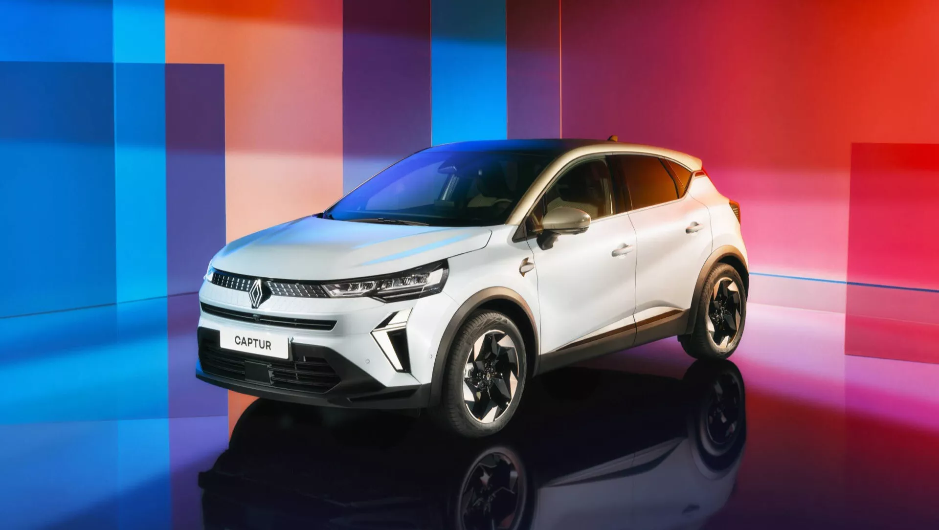 Facelift Renault Captur II 2024 v ateliéru, pohled z nízkého úhlu zepředu a ze strany zdůrazňující jeho moderní design kompaktního SUV na barevném abstraktním pozadí přecházejícím od modré po růžovou, zvýrazňuje přední mřížku, LED světlomety, elegantní nárazník, sochařsky zpracovanou kapotu, blatník a sportovní slitinová kola.
