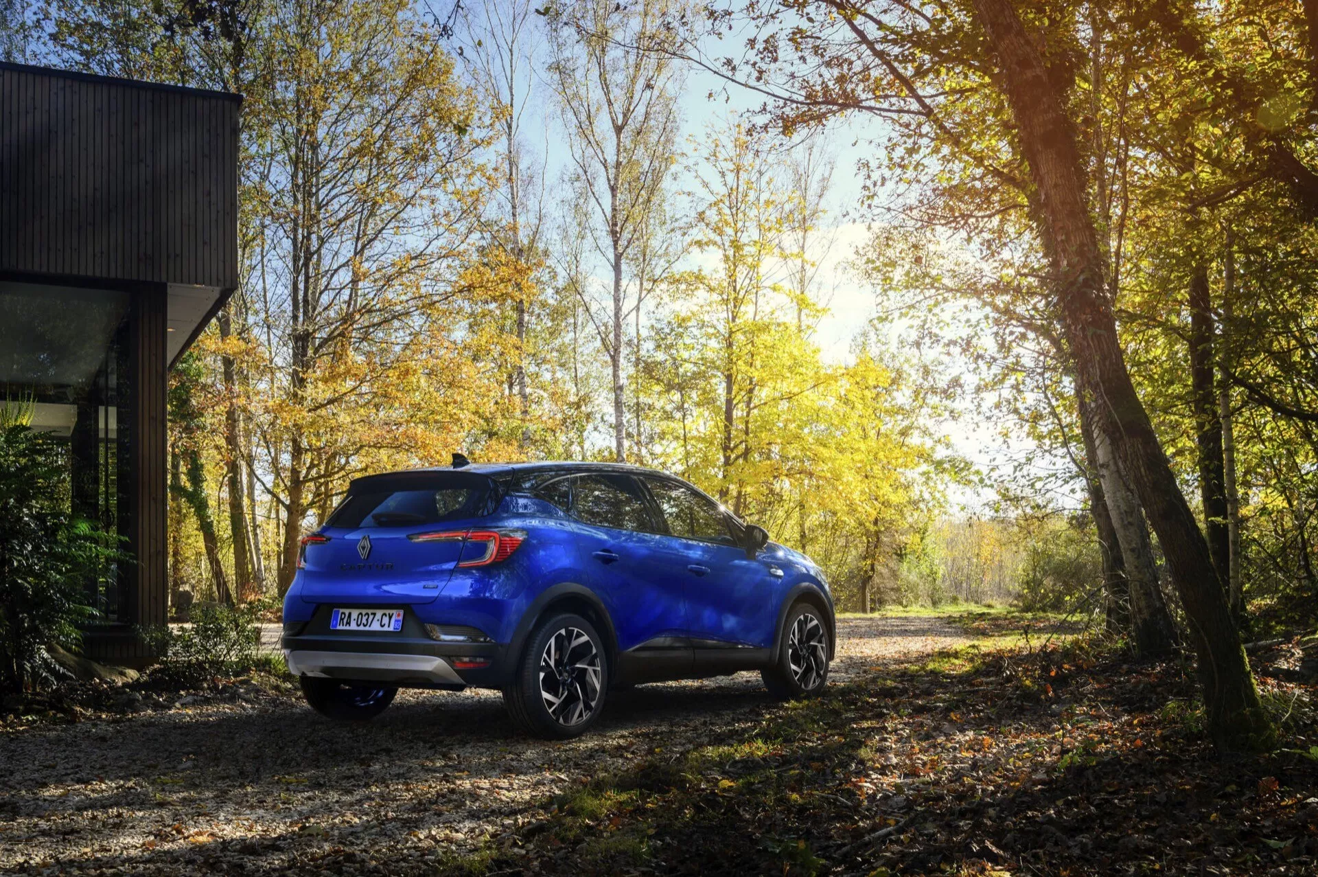 Modré kompaktní SUV Renault Captur II Facelift ročník 2024 zaparkované venku na nezpevněné cestě, předvádějící zadní a boční profily s LED zadními světly, slitinovými koly, parkovacími senzory a zavěšením pro lehkou terénní jízdu v klidné lesní oblasti v podzimním období se slunečními paprsky procházejícími mezi stromy.