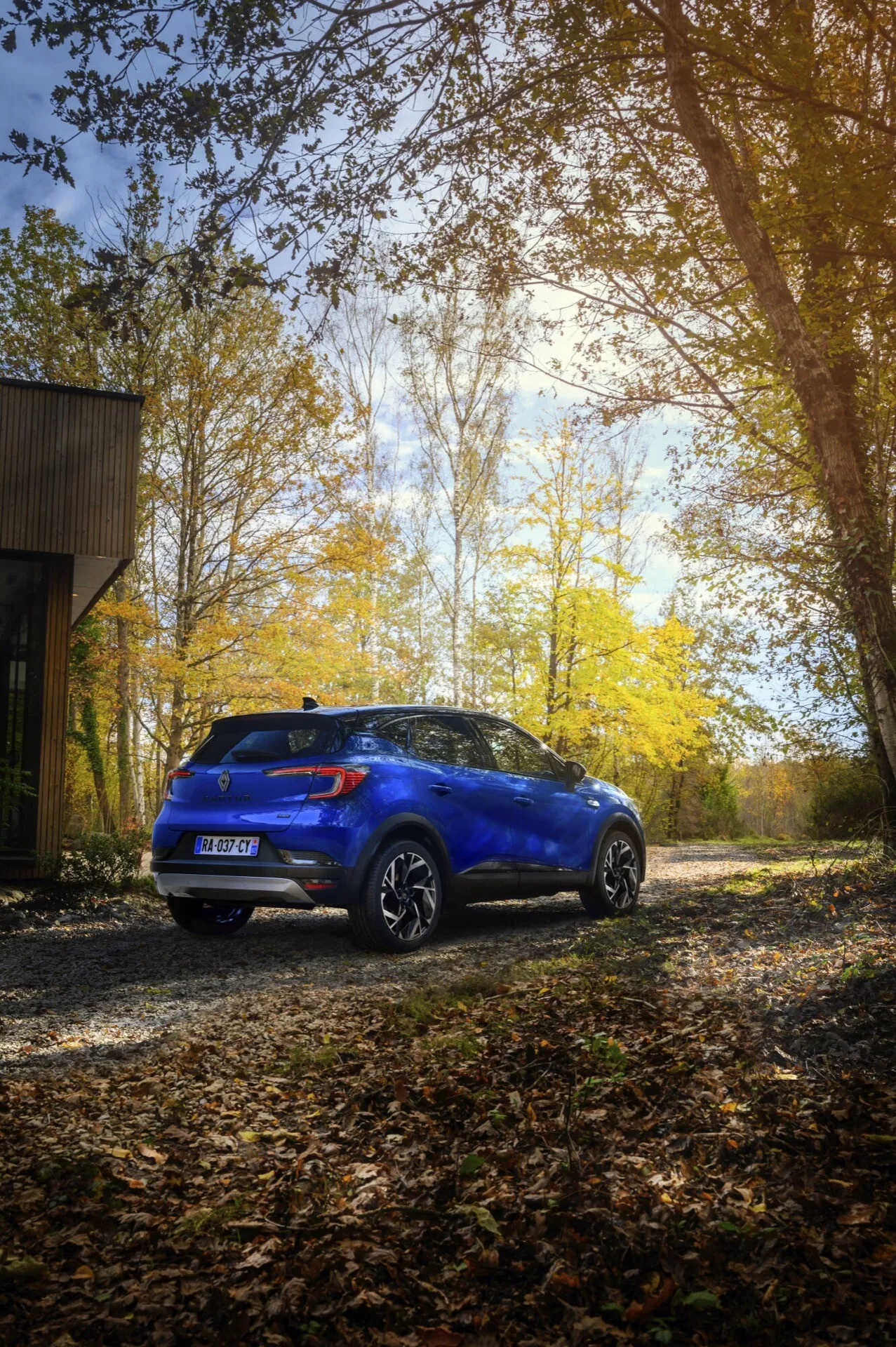 Modrý model Renault Captur II facelift 2024, který zvenku na štěrkové ploše s podzimním lesním pozadím ukazuje svůj zadní a boční profil, zdůrazňuje jeho designové linie, lesklý povrch, slitinová kola a zadní spoiler.
