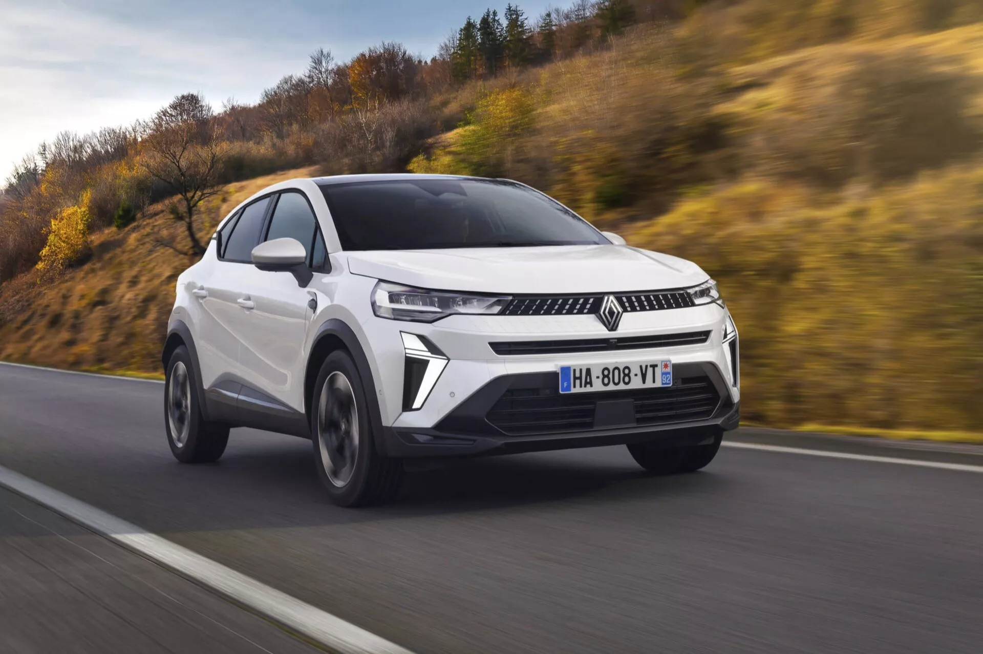 Renault Captur 2024 (II-facelift) ve dynamickém tříčtvrtečním předním pohledu zdůrazňujícím jeho elegantní crossoverový design, charakteristickou mřížku chladiče, ostrá LED denní světla, moderní jednotky světlometů, aerodynamický přední nárazník s nasávacími otvory, sochařsky vypracované linie kapoty, slitinová kola s více paprsky, zrcátka s integrovanými směrovými signály, metalický lak a panoramatické střešní okno na pozadí s rozmazaným pohybem, což ztělesňuje esenci moderní automobilové technologie a designové estetiky.