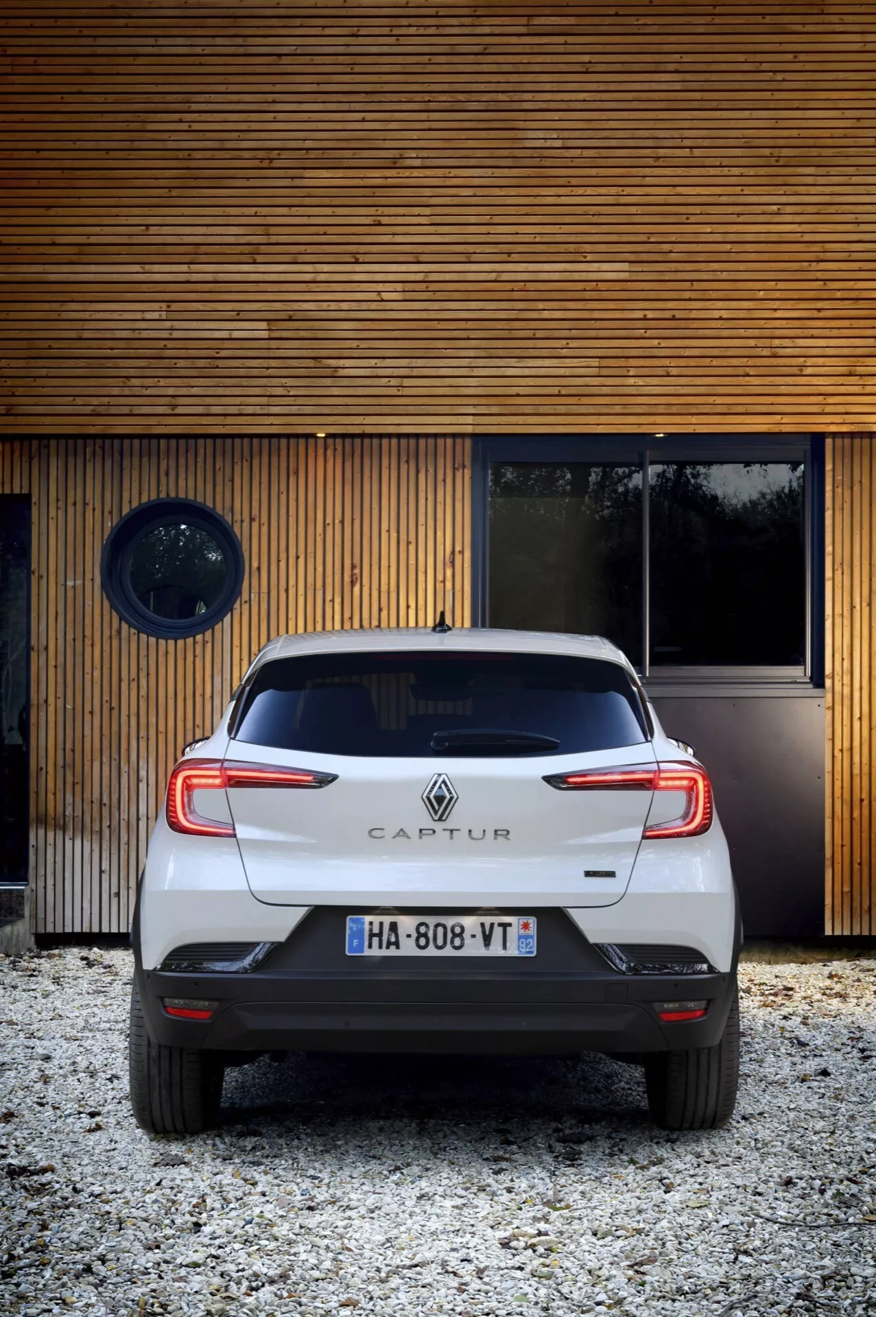 Zadní pohled na Renault Captur 2024 (II facelift), kompaktní crossover, ukazující zadní světla, nárazník, dveře kufru a registrační značku z úhlu zdola nahoru, s pozadím moderní struktury a osvětlením denního světla