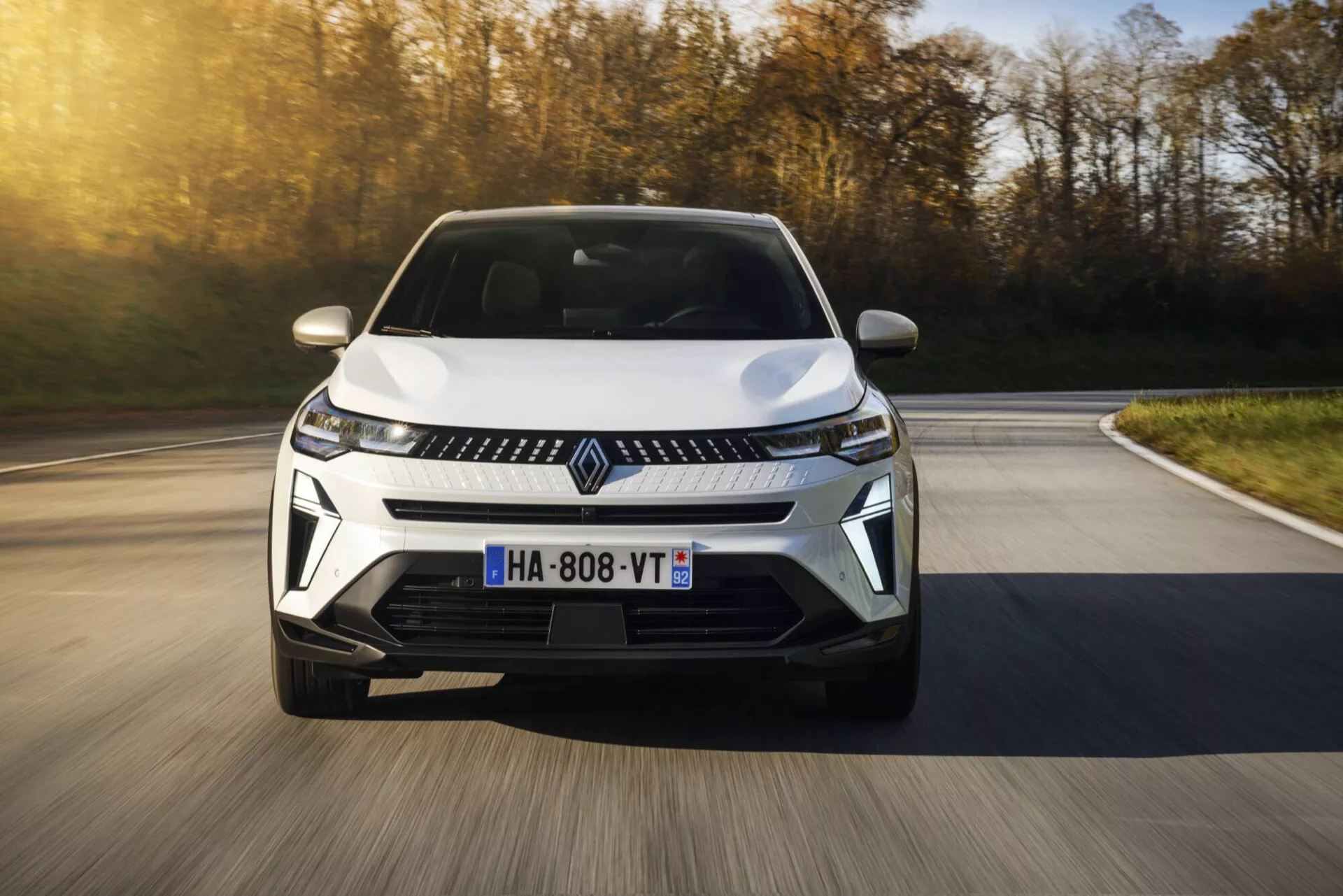 Facelift Renault Captur 2024 v pohybu z nízkého úhlu pohledu zepředu, zdůrazňující jeho dynamický design a moderní prvky včetně LED světlometů, elegantní mřížky s centrálním znakem a aerodynamického předního nárazníku na pozadí s rozmazaným podzimním landschaftem