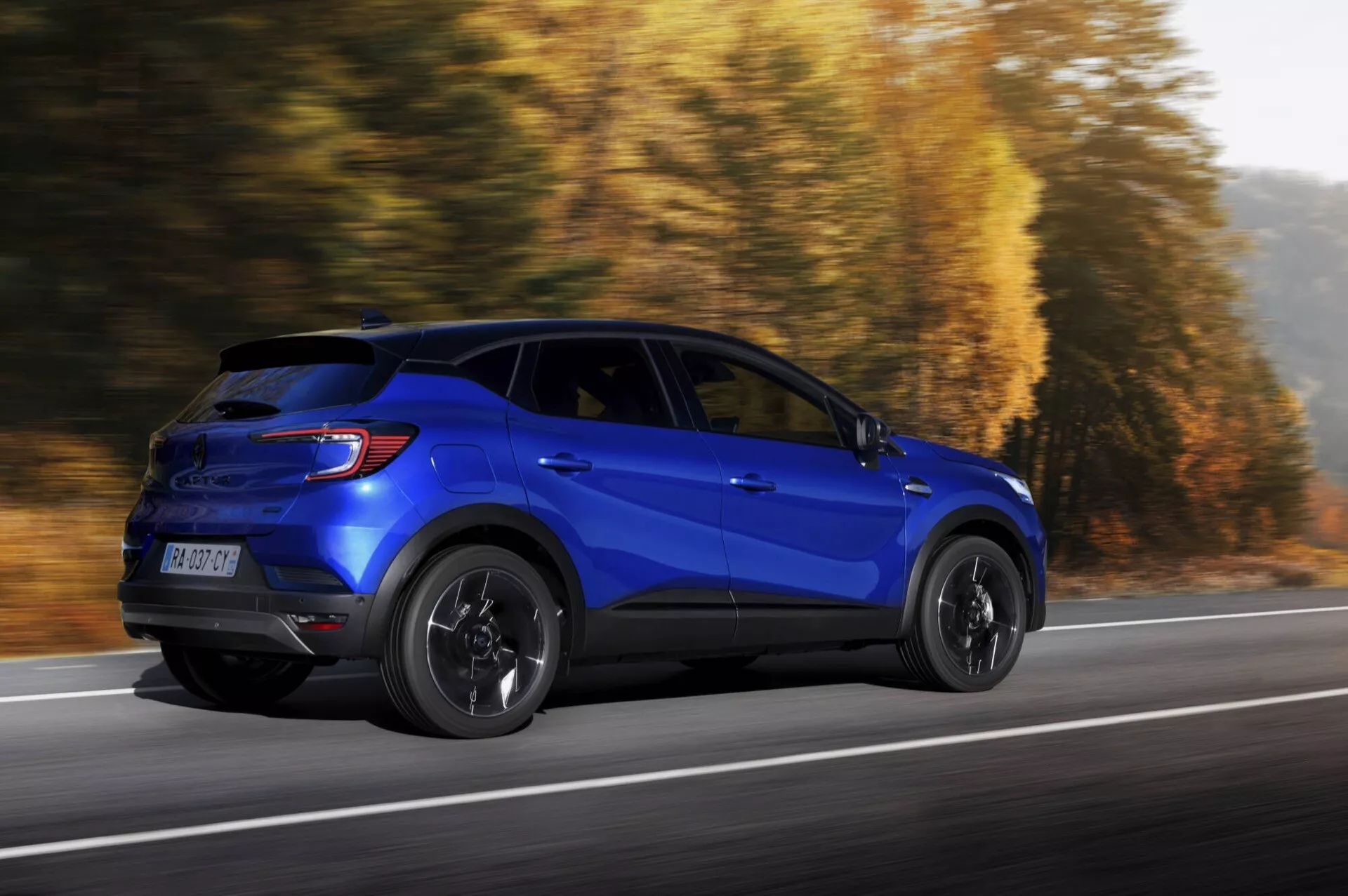 Modré sportovní užitkové vozidlo Renault Captur 2024 (II facelift) v dynamickém pohybu, profilový pohled zdůrazňující stranu a zadní designové prvky na silnici s podzimním pozadím