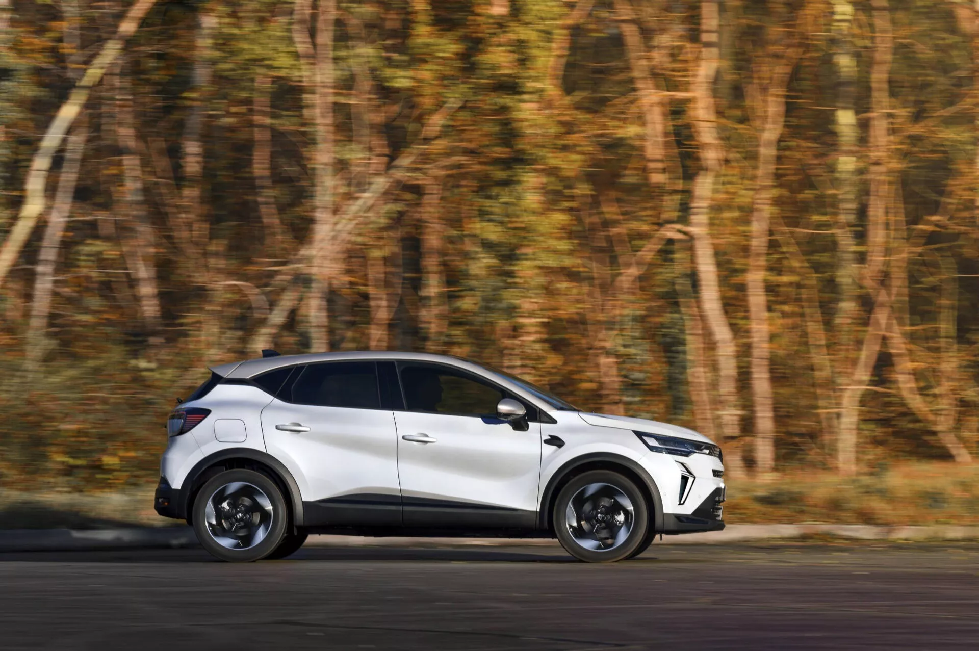 Renault Captur 2024 po faceliftu v pohybu: moderní bílý crossover s detailním pohledem ze strany a úhlem zezadu na rozostřeném podzimním lesním pozadí