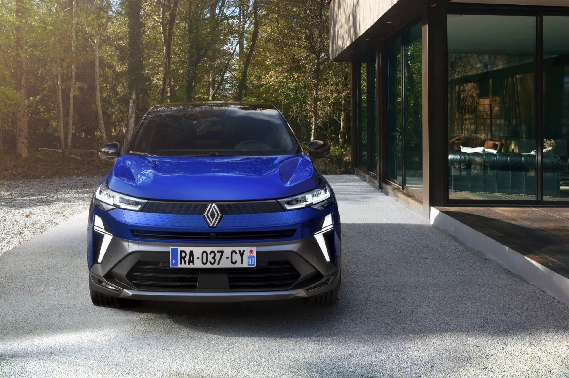 Přední pohled na Renault Captur II Facelift 2024 zdůrazňující elegantní světlomety, moderní mřížku chladiče, živou modrou barvu a sportovní design nárazníku s integrovanými větracími otvory, zaparkovaný na příjezdové cestě s pozadím moderního domu