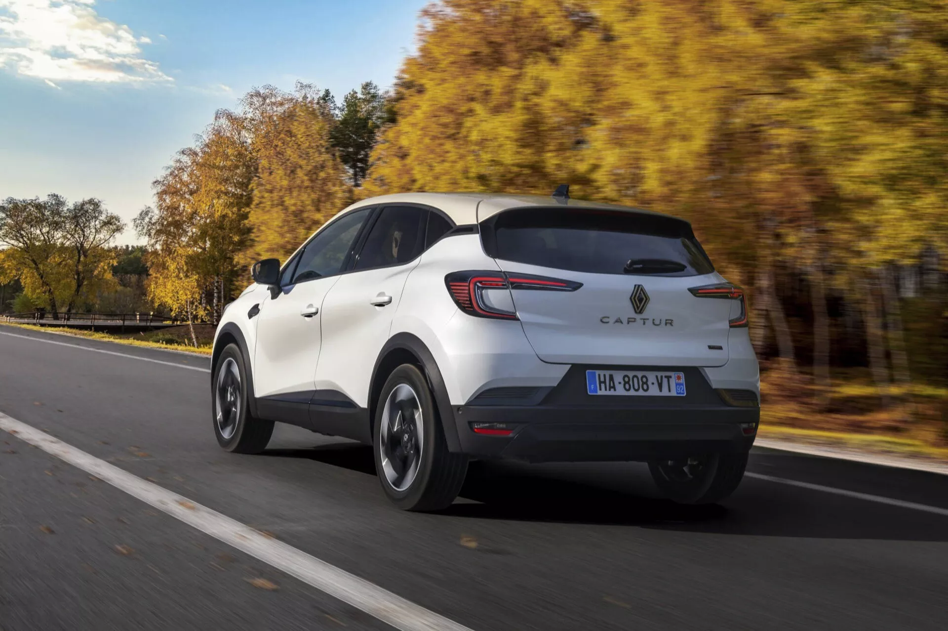 Dynamický pohled zezadu na tři čtvrtiny Renault Captur 2024 (facelift II) v bílé barvě, který prezentuje designové prvky jako zadní světla, tvar zadních dveří a nárazník na silnici lemované stromy s podzimním listím