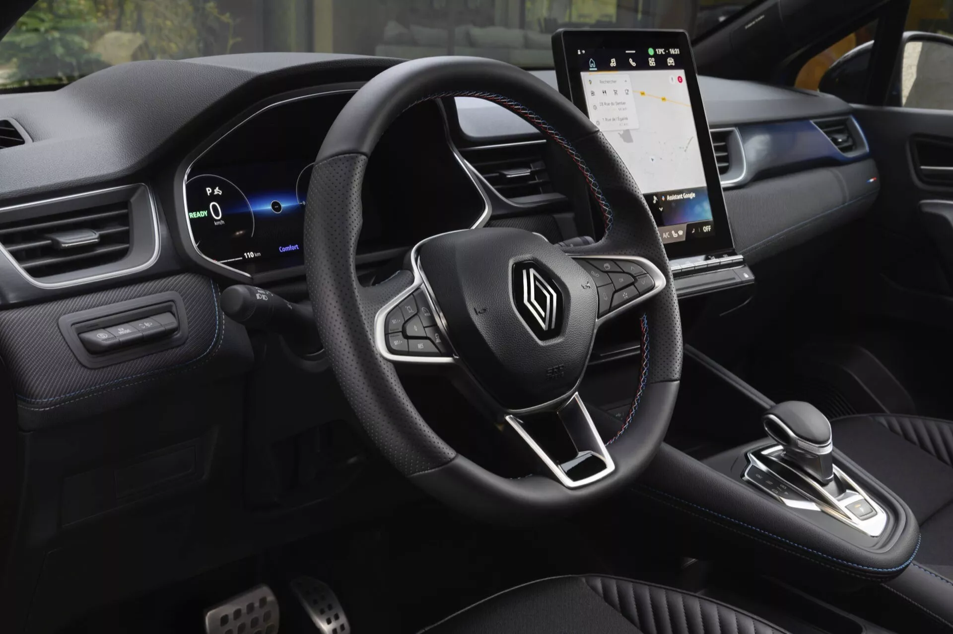 Interiér Renault Captur II facelift 2024 z pohledu spolujezdce vpředu, ukazující multifunkční volant s logem, digitální palubní desku, dotykový informační systém, řadicí páku pro automatickou převodovku, palubní desku ve dvoutónovém provedení s měkkými materiály a dekorativním prošíváním a sportovní pedály, což zdůrazňuje moderní design zaměřený na pohodlí, komfort a technologie.