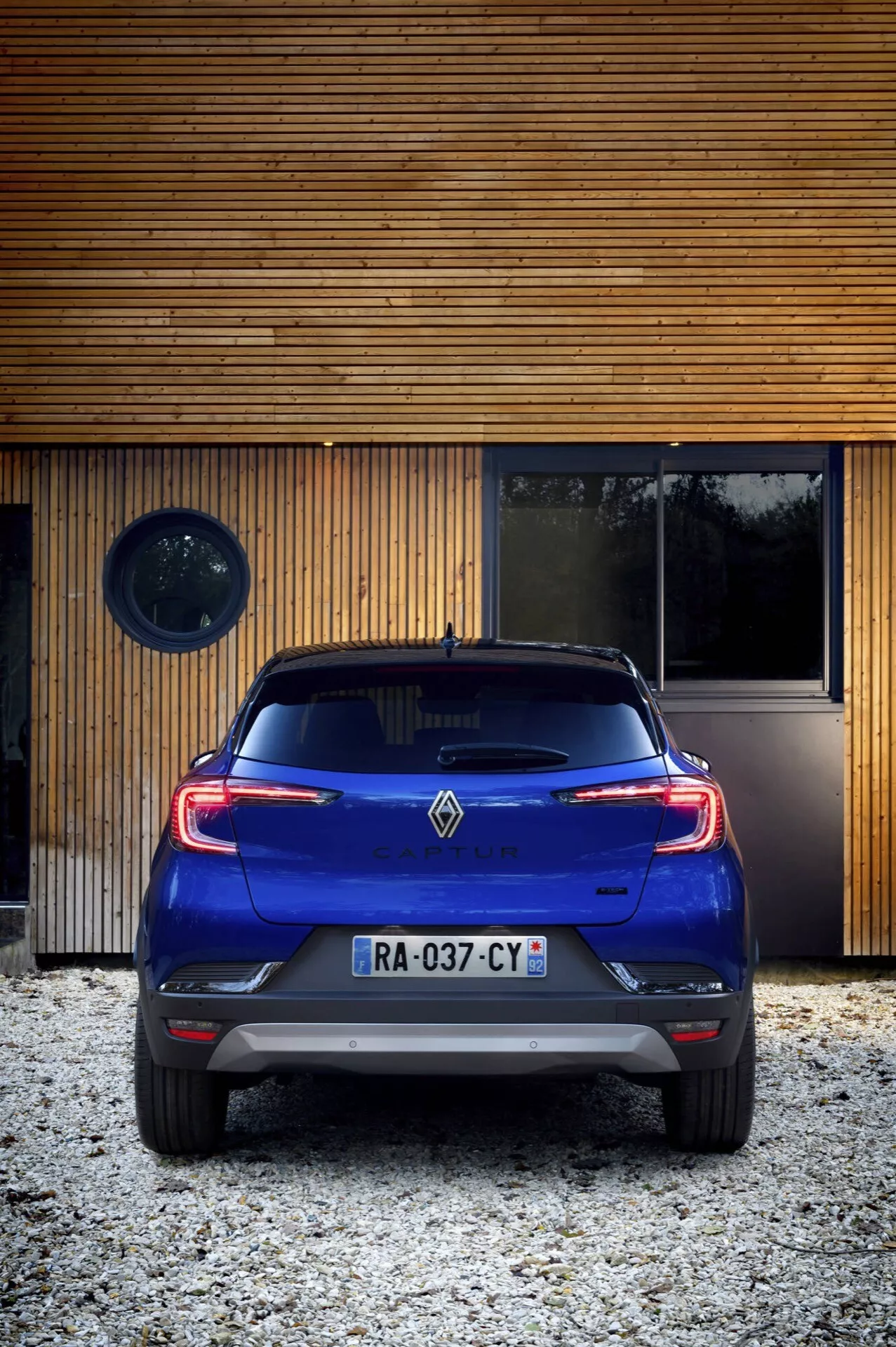 Zadní pohled na Renault Captur 2024 (II Facelift) na štěrku s moderní dřevěnou konstrukcí na pozadí, předvádějící designové prvky jako jsou zadní světla, zadní nárazník, logo a charakteristické zadní čelní sklo