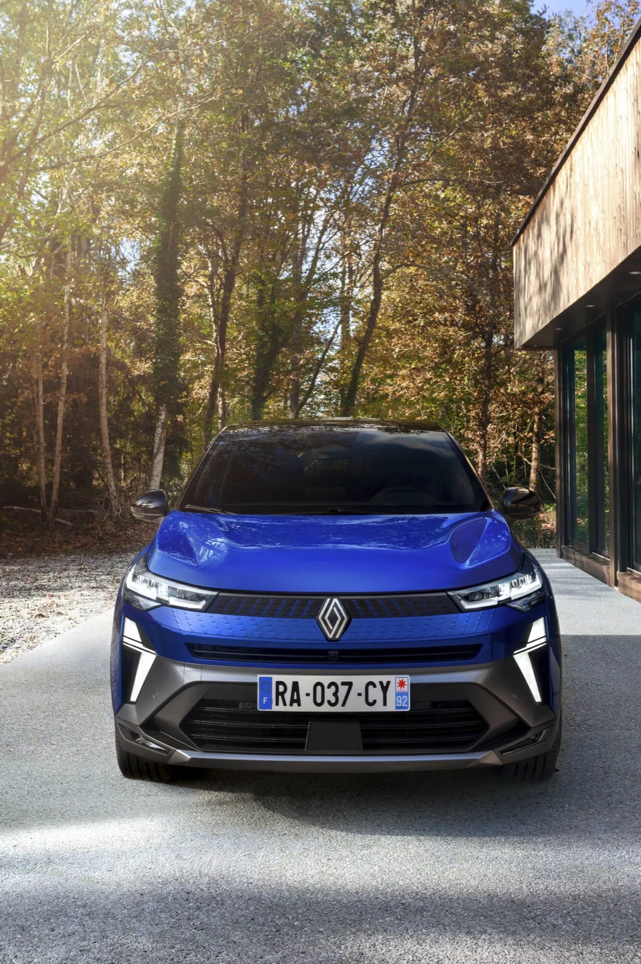 Pohled zepředu na facelift Renault Captur 2024 ukazující design mřížky, LED světlomety a emblém na asfaltové příjezdové cestě s dřevinami a budovou s velkými skleněnými okny na pozadí v slunečných podmínkách.