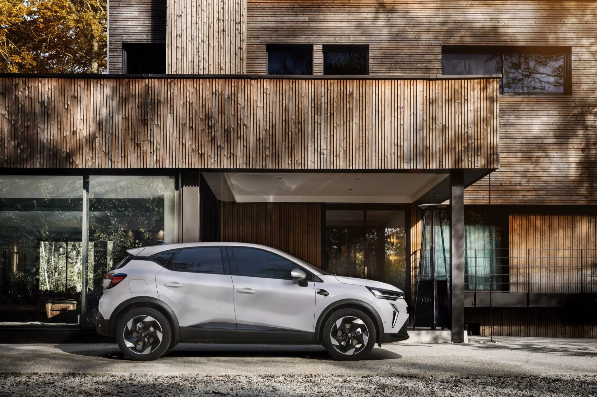 Boční profil Renault Captur II facelift 2024 parkovaný před moderním domem se dřevěnou fasádou a velkými skleněnými okny, stylová černá slitinová kola, elegantní design SUV se čtyřmi dveřmi a aerodynamická zrcátka
