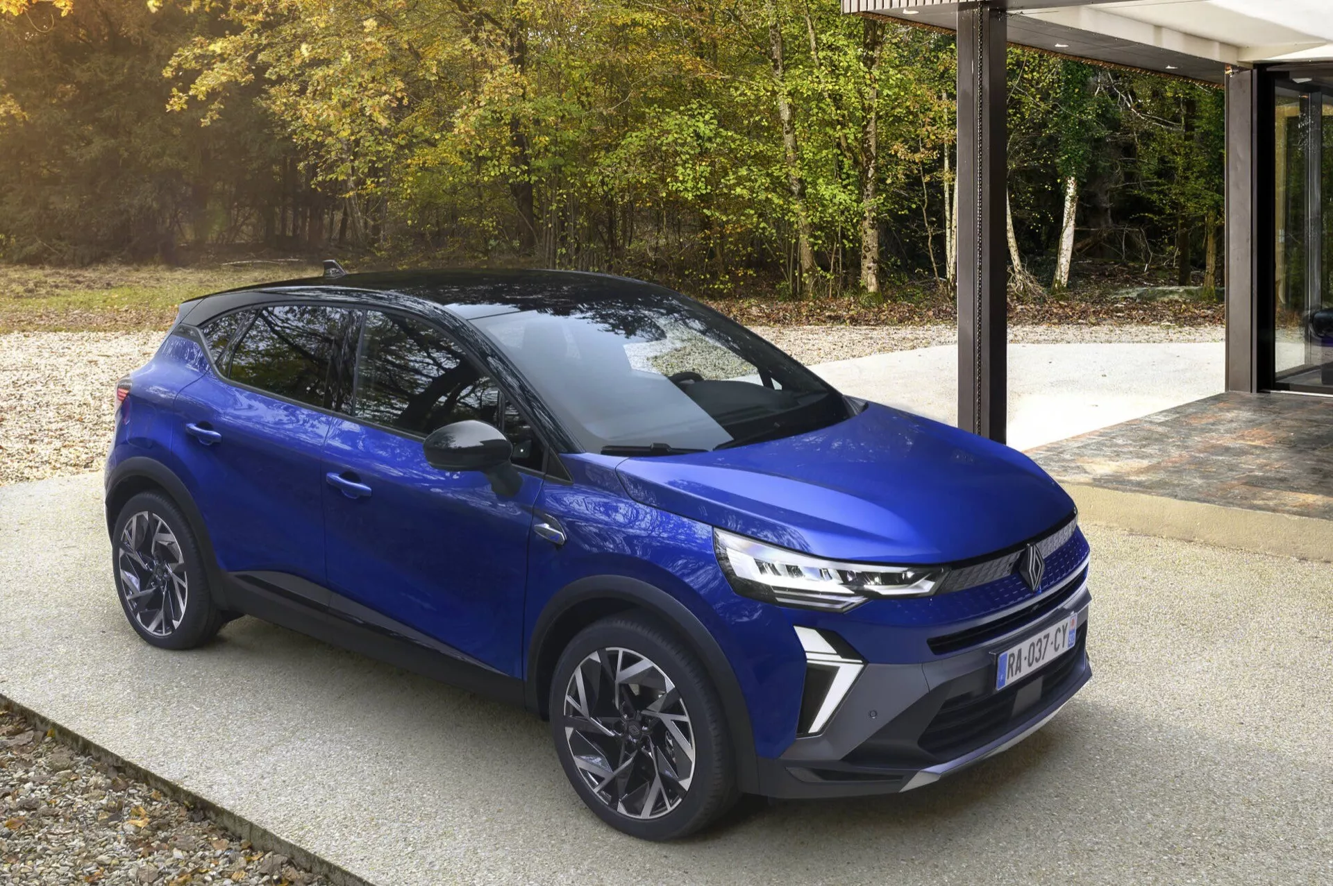 Renault Captur (II-Facelift) 2024 v moderní modré barvě, zaparkované na příjezdové cestě s přírodním pozadím stromů a moderní architekturou, předvádějící přední mřížku, LED světlomety, slitinová kola a boční profil ve dne.