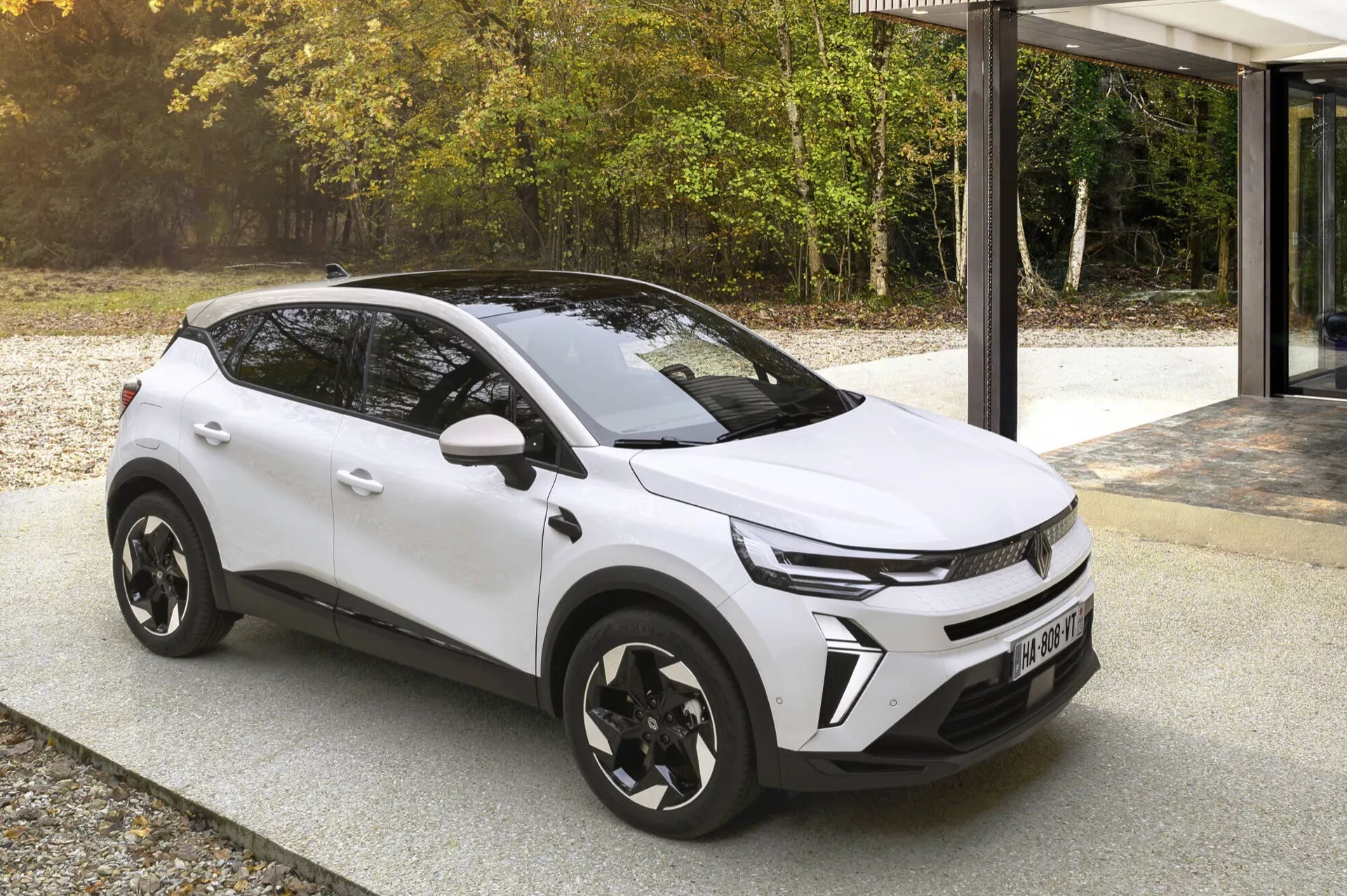 Facelift Renault Captur II 2024 v bílé barvě, ukazující jeho dynamickou přední mřížku, LED světlomety a aerodynamický design s černými oblouky kol a výrazným designem slitinových kol, zaparkovaný na vydlážděné ploše u moderní budovy se skleněnou fasádou