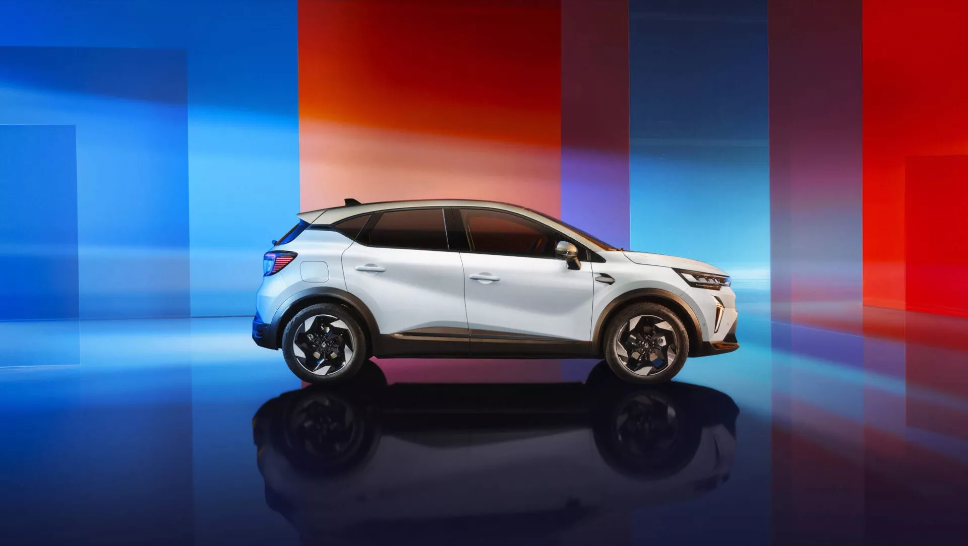 Renault Captur II facelift 2024 fotografovaný z profilu na pozadí moderního přechodu z modré do červené, představující jeho aerodynamická zpětná zrcátka, zapuštěné kličky dveří, složité slitinové kola, aerodynamická okna a sportovní linii střechy.
