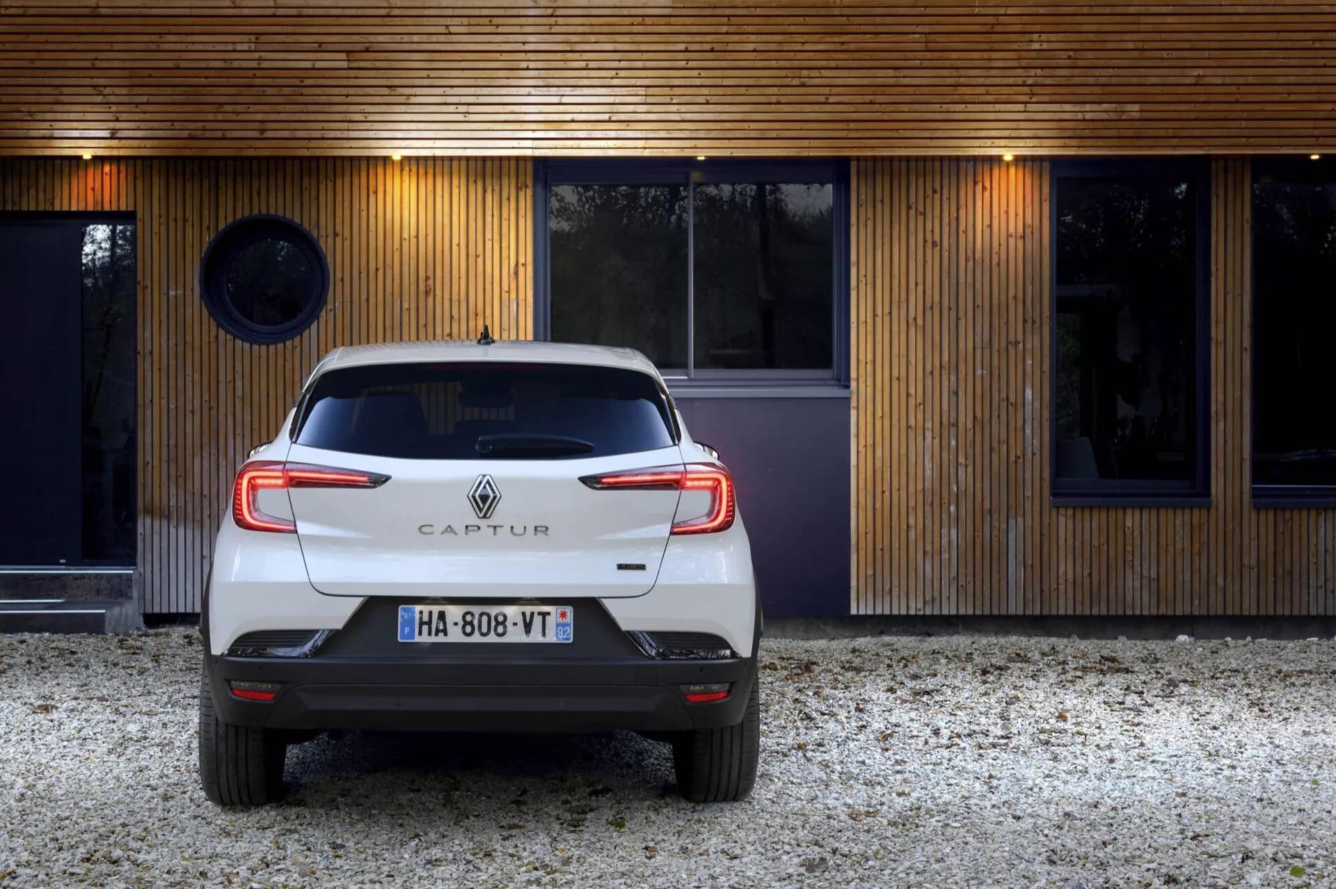 Zadní pohled na bílý Renault Captur 2024 (II-facelift) zaparkovaný před moderní budovou se dřevěnou fasádou, ukazující C-tvarovaná LED zadní světla, emblém a parkovací senzory.