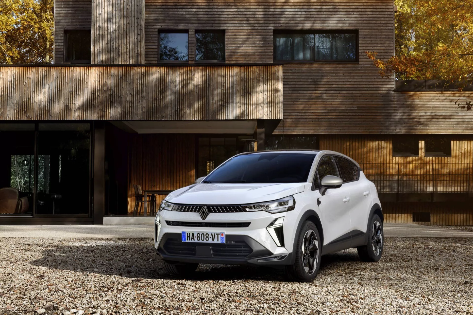 Facelift model Renault Captur 2024 zaparkovaný pod úhlem předvádějící moderní design SUV, s výraznou přední mřížkou s horizontálními liniemi, štíhlými světlomety s LED denními světly, aerodynamicky tvarovanou kapotou a sportovním skloněným střešním profilem na pozadí luxusního rezidenčního domu