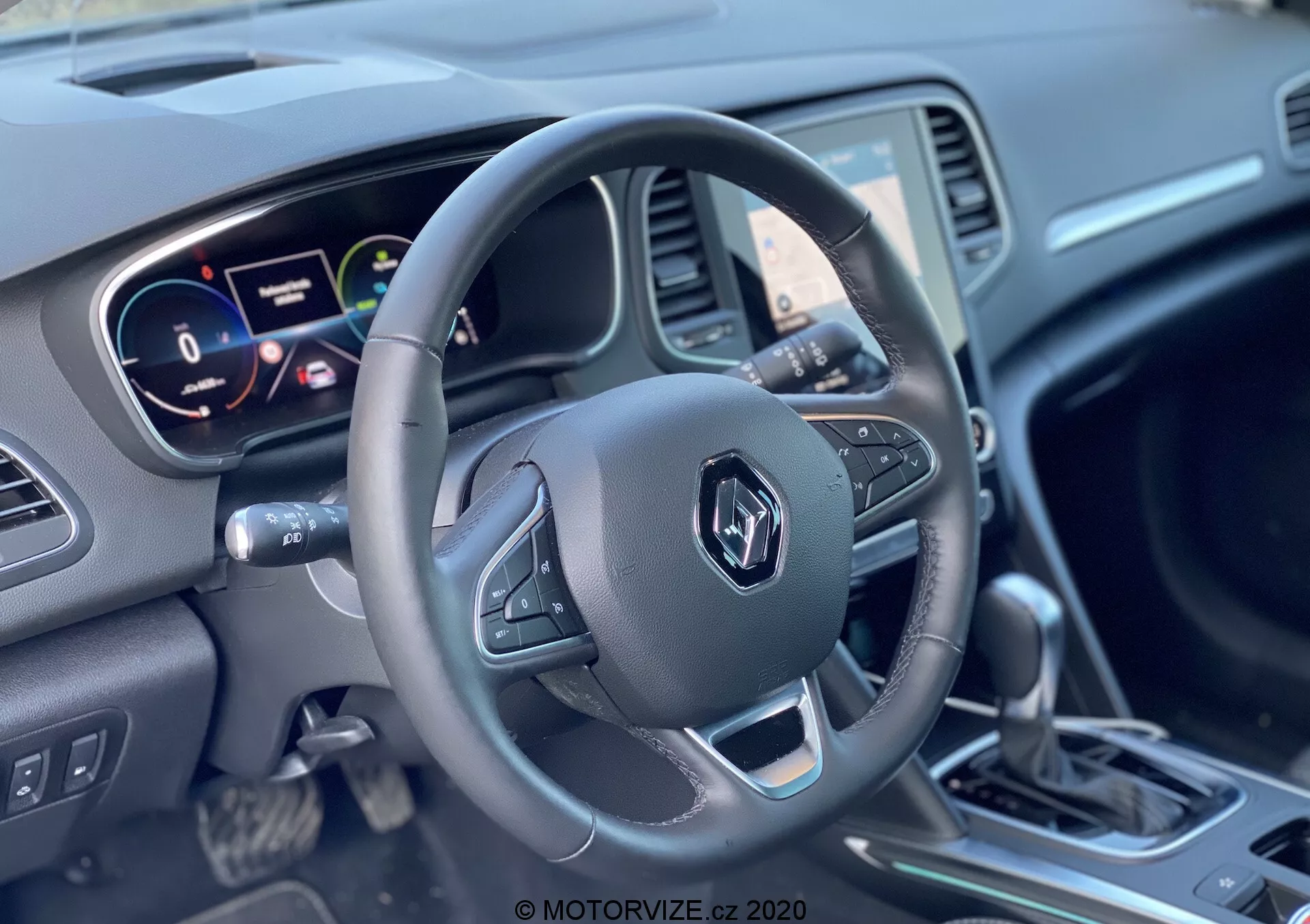 Pohled na interiér Renault Megane E-Tech 2022 zaměřený na pohled řidiče na volant a přístrojovou desku, zobrazující tříramenný volant s ovládacími tlačítky, digitální a analogový přístrojový panel a řadicí páku obklopenou různými ovládacími prvky.
