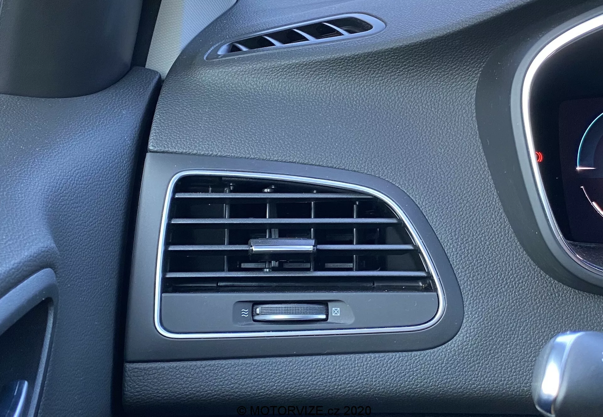 Detailní pohled na větrací otvor a ovládací knoflík klimatizace na straně spolujezdce v Renaultu Megane 2022 E-Tech. Černé lamely větráku jsou orientovány horizontálně, s malým knoflíkem na vertikální ose pro nastavování proudění vzduchu. Malá část neidentifikovaného přístroje nebo panelu je trochu viditelná napravo. Povrchy palubní desky mají texturovaný černý plastový povrch osvětlovaný okolním světlem.