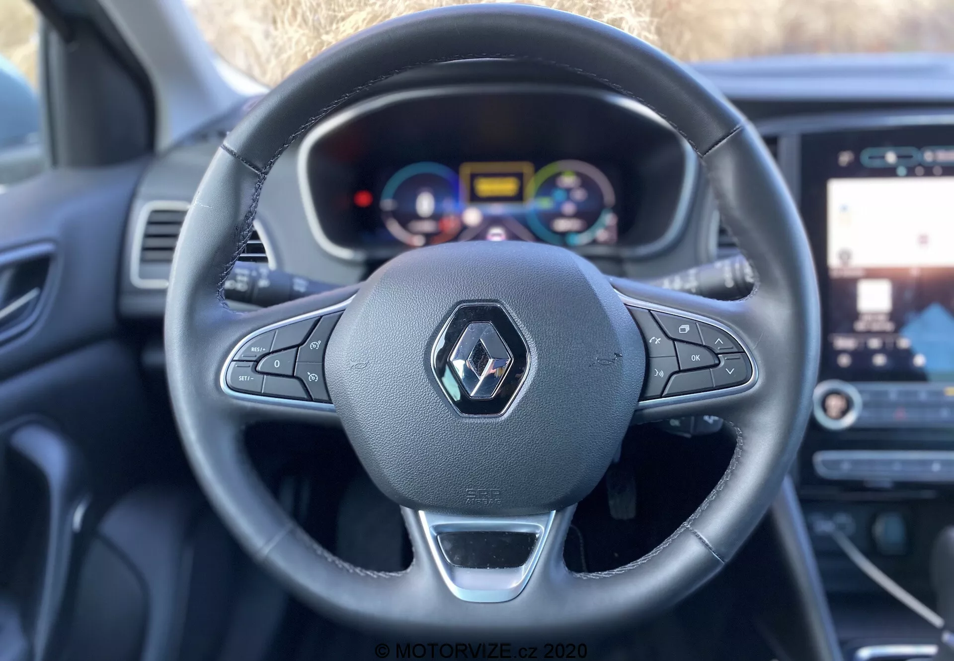 Pohled na interiér Renaultu Megane E-Tech 2022 z perspektivy řidiče, ukazující tříramenný volant s ovládacími prvky a kovovým akcentem, přizpůsobitelný digitální přístrojový panel s jasně barevnými ukazateli a indikátory asistenčních funkcí řidiče, pohled na dotykový infotainment se zobrazením map/navigace a moderní, ergonomický interiér s koženými nebo podobnými materiály a syntetikou, foceno z pohledu řidiče s mělkou hloubkou ostrosti zaměřující se na volant a palubní desku.