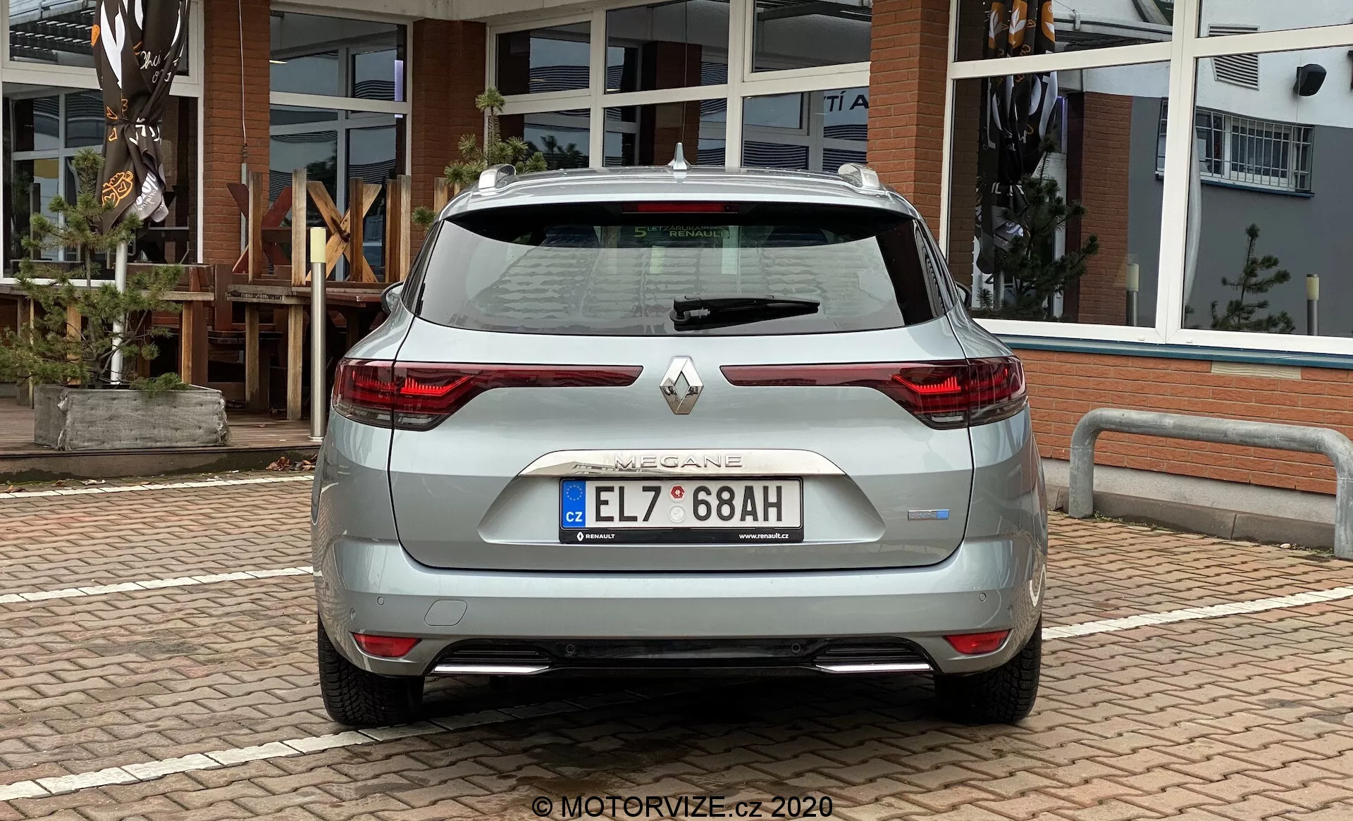 Zadní pohled na světlý hatchback vyrobený společností Renault, který má na celé šířce zadní vrata s integrovaným spojlerem, moderní LED zadní světla ve tvaru C, parkovací senzory, zpětnou kameru, dvojitý výfukový systém a chromové lišty na zadním nárazníku. Vozidlo je zaparkováno venku na dlážděné ploše s budovou se skleněnými dveřmi a okny v pozadí.
